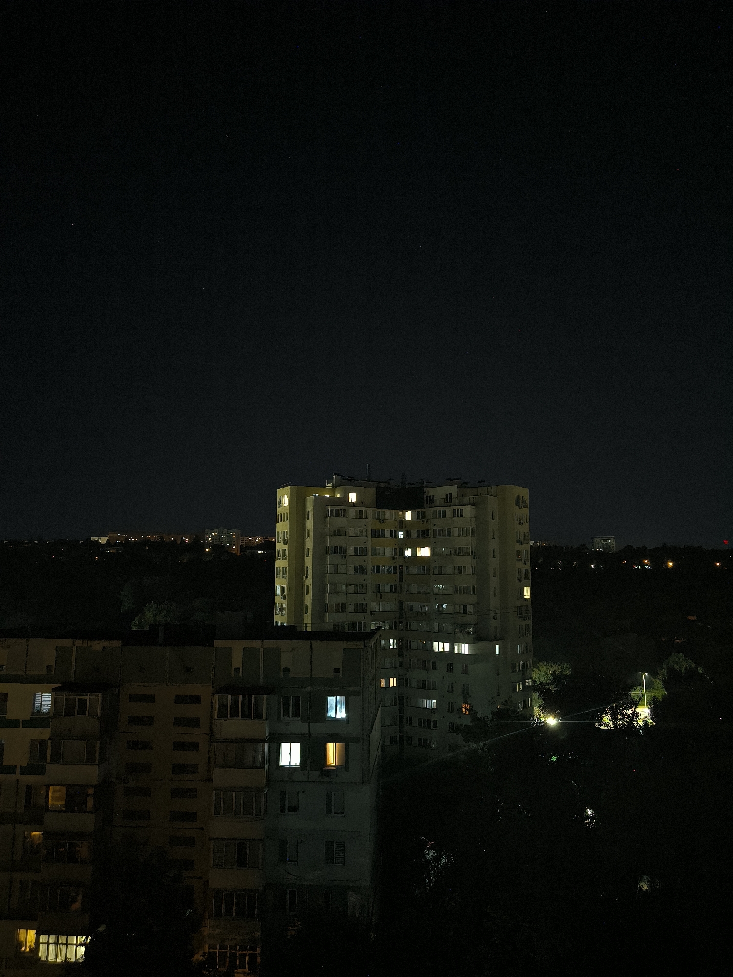 This is a night photograph taken from a high vantage point, likely an apartment building, overlooking a residential area in Chișinău, Moldova. The scene is dominated by the dark night sky and silhouetted trees. In the mid-ground, two apartment buildings are visible. The building on the right is a taller, pale yellow structure with many lit windows, suggesting occupancy. It has a distinctive, somewhat dated architectural style common in post-Soviet urban planning. To the left and slightly closer to the foreground is another, darker apartment building, appearing less lit than the one on the right, with some windows showing dim yellow light. The background shows a distant cityscape with scattered lights. A street lamp is visible in the middle ground, illuminating a patch of trees. The overall impression is one of urban nighttime quietude.
