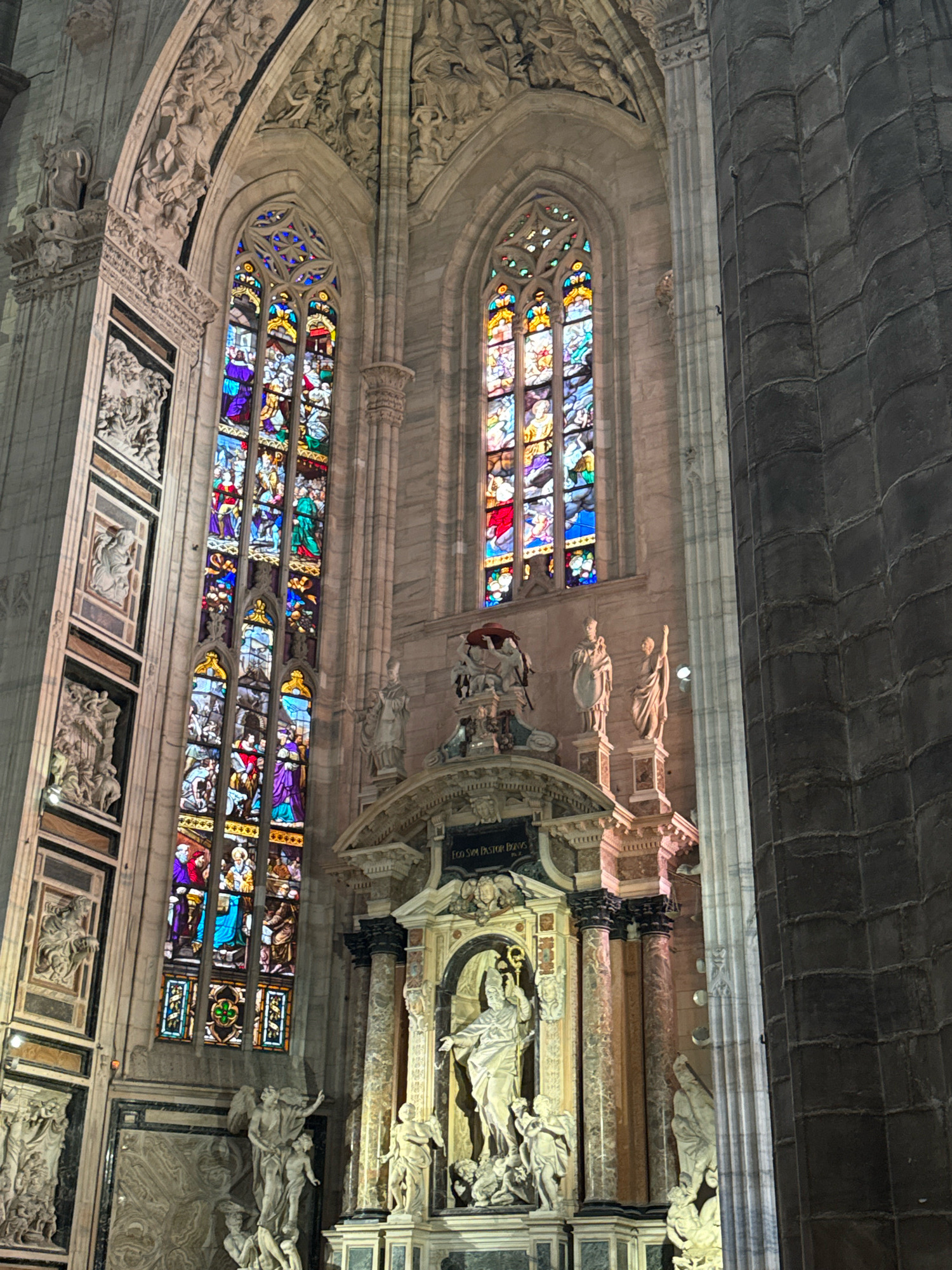This image depicts the interior of the Milan Cathedral (Duomo di Milano) in Milan, Italy. The photograph focuses on a section of the cathedral featuring large, ornate stained-glass windows that are brightly colored and depict various scenes. Below the windows, a detailed altar or monument is visible, adorned with sculptures of figures and architectural elements. The cathedral's interior is characterized by grand columns, intricate stonework, and a high vaulted ceiling, typical of Gothic architecture. The lighting suggests an indoor environment, possibly with some natural light filtering through the windows.
