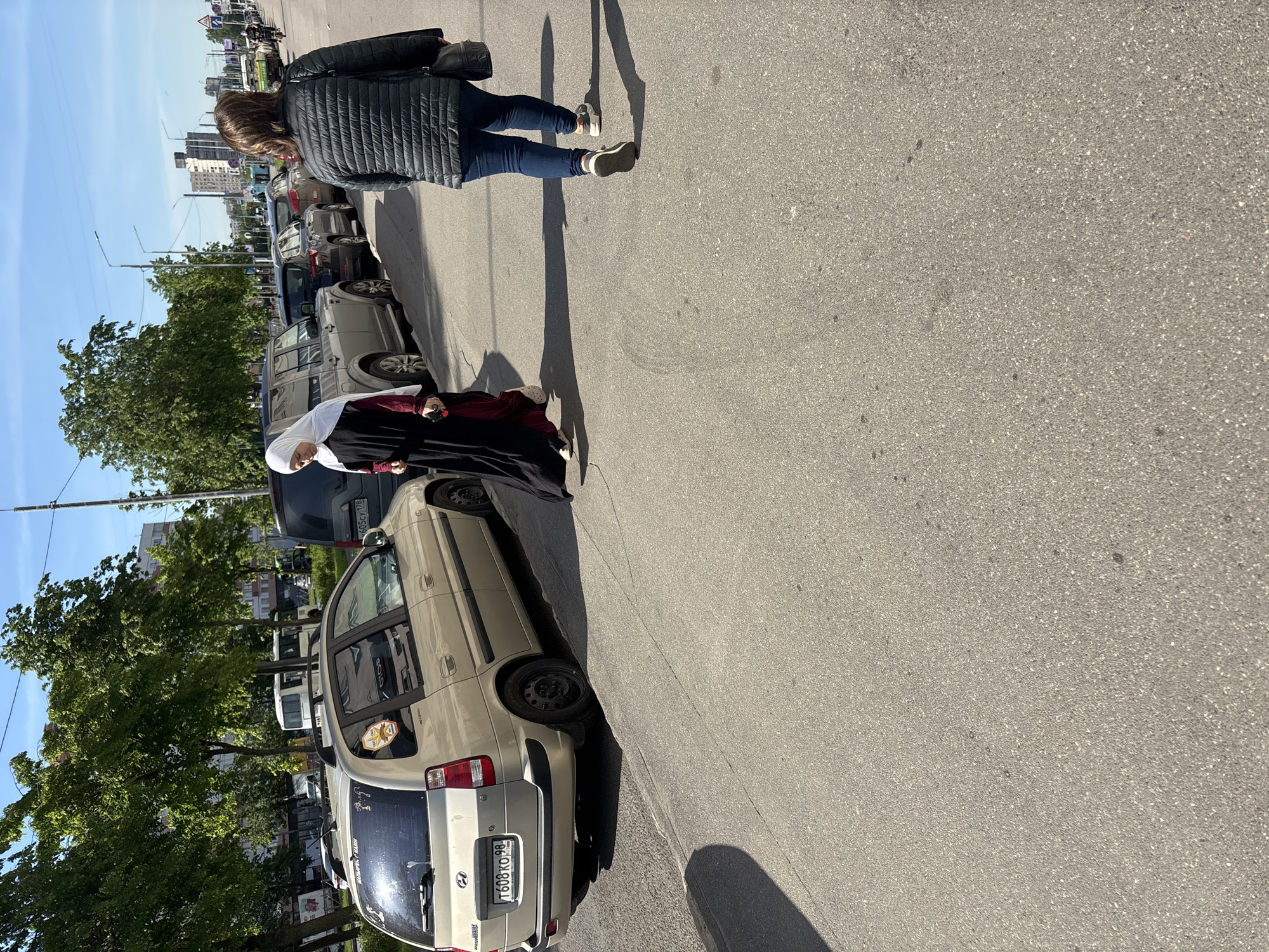 The image depicts an outdoor urban scene in Russia, likely Saint Petersburg, on a sunny day with clear blue skies. The street is paved with asphalt and lined with parked cars on one side, while trees with green foliage are visible behind them. Buildings can be seen in the distance, suggesting an urban or suburban environment.
Two women are visible on the right side of the frame, walking or standing alongside the paved road. The woman in the foreground is seen from behind, wearing a dark puffer jacket, blue jeans, and light-colored sneakers. She casts a long shadow on the asphalt, indicating either morning or late afternoon sunlight. Ahead of her, closer to the parked vehicles, is another woman wearing a white head covering (hijab), a dark long dress or abaya, and a red undergarment or accent piece. She appears to be looking towards the parked cars.
A prominent light-gold/beige Hyundai car, possibly a Starex or similar model, is parked in the mid-ground. Its rear license plate, `T608KO 98`, identifies it as being from the Saint Petersburg region of Russia. Several stickers adorn its rear window, including one resembling a sheriff's badge and some barely legible text. Other cars are parked further down the street. Utility poles or streetlights are also visible along the roadside. The overall impression is that of everyday street activity under good weather conditions.