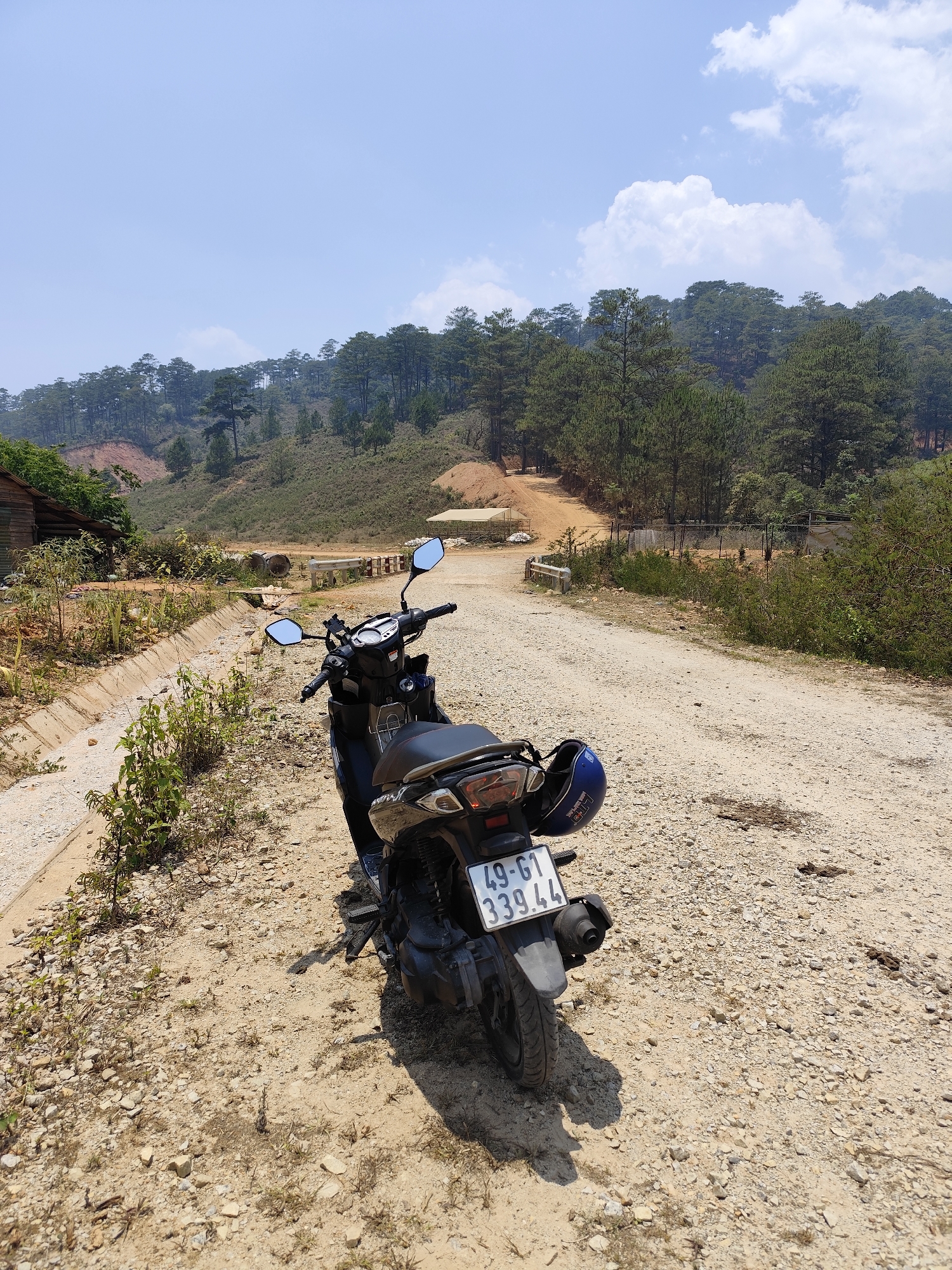 The scene is set on a dirt road in Lạc Dương, Vietnam, under a blue sky with scattered clouds. A black scooter with a blue helmet strapped to the back is parked on the road. The scooter's license plate reads "49-G1 339.44". The road leads towards a small bridge and a hillside covered with trees. To the left of the scooter is a ditch with vegetation, and a small wooden structure is visible in the background. The road surface is gravel and dirt.