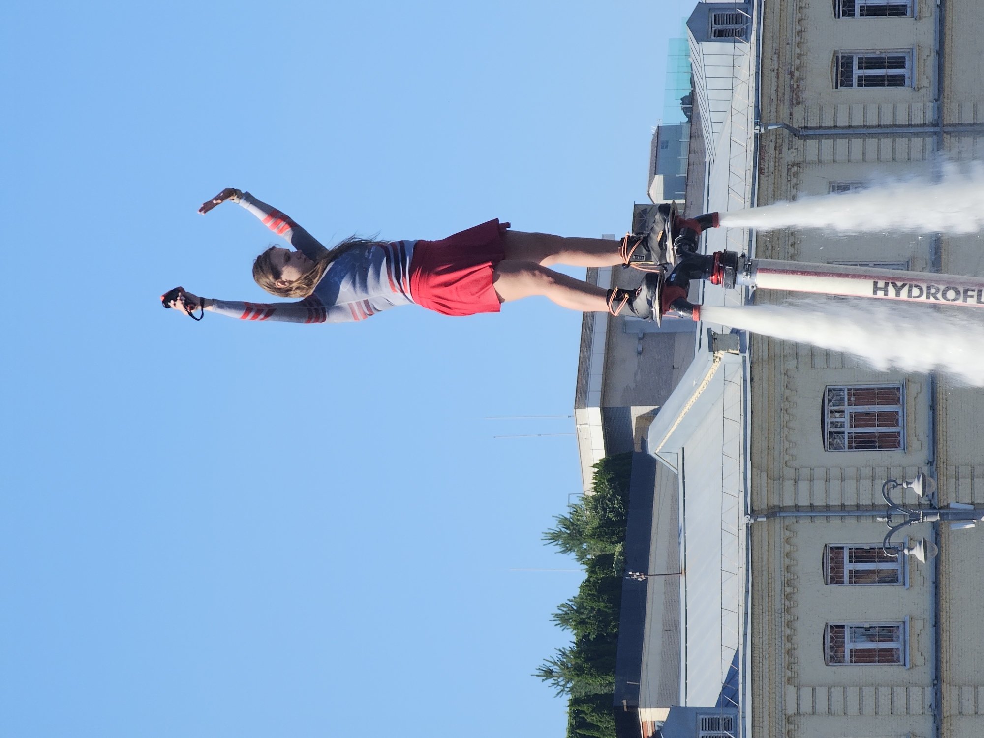 A person is performing on a flyboard over the Moskva River in Moscow, Russia. The person is wearing a grey long-sleeved shirt with red stripes and a red skirt. They are holding onto a controller and have their arms raised in the air. The flyboard is attached to their feet and is spraying water upwards. In the background, there are buildings and trees. The sky is blue.