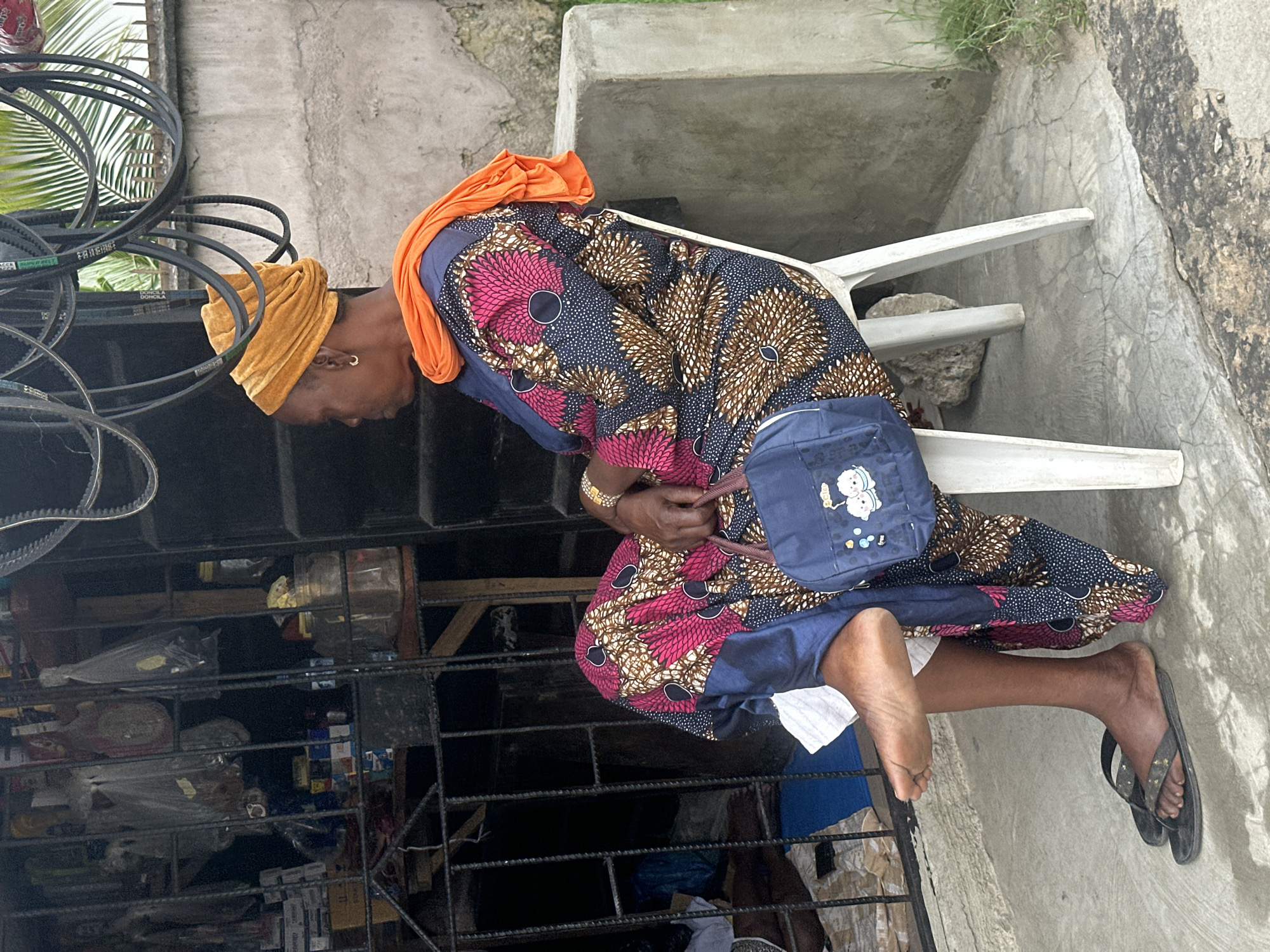 An elderly African woman sits on a white plastic chair outside a shop in Badagry, Nigeria. She is wearing a bright orange headwrap, a colorful patterned dress with pink, brown, and dark blue motifs, and an orange scarf draped over her shoulders. A small blue purse with cartoon characters is held in her right hand. Her left foot is bare and propped up, while her right foot wears a black flip-flop. The shop behind her is stocked with various items, including bottles, jars, and packaged goods, visible through a metal grate. Overhead, several black belts are suspended. The ground is concrete, and a rough concrete wall with some grass growing at its base is to the right of the woman.