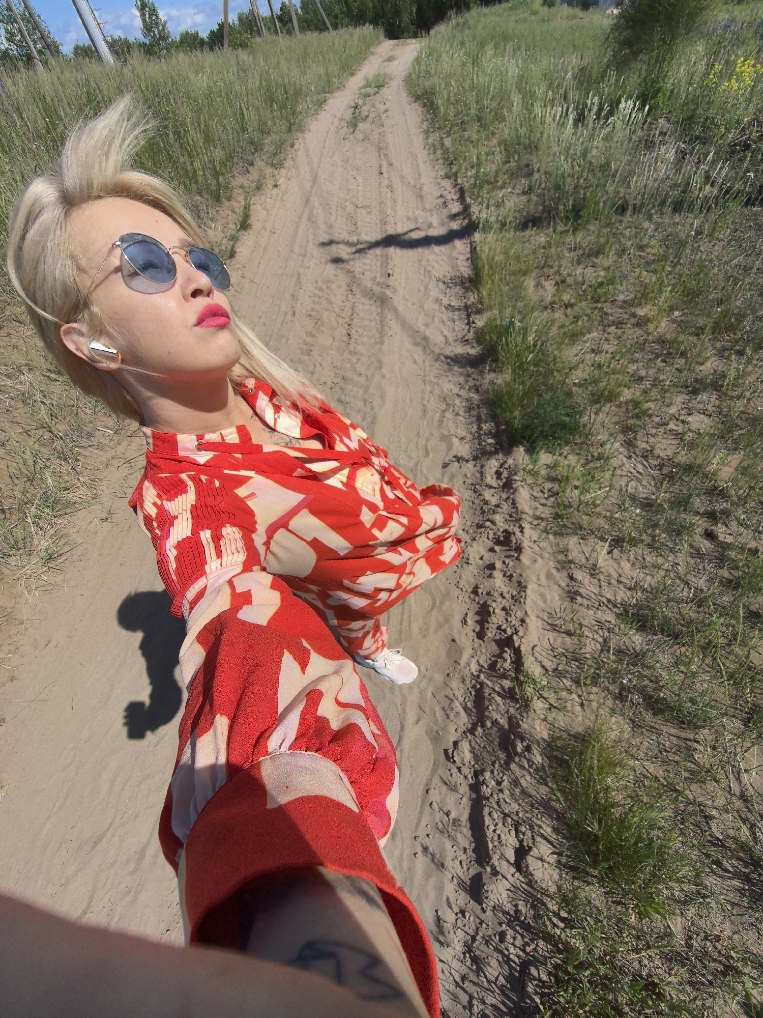 A blonde woman with bright pink lipstick and blue sunglasses is captured in a selfie while walking on a dirt path in Noviy, Russia. She is wearing a red and white patterned shirt with voluminous sleeves and white sneakers. In her left ear, she has a white wireless earbud. The path is surrounded by tall, dry grass and sparse trees. The lighting suggests it is daytime, with strong sunlight casting shadows. The image appears to be taken in a rural or natural outdoor setting within the city limits of Noviy.
