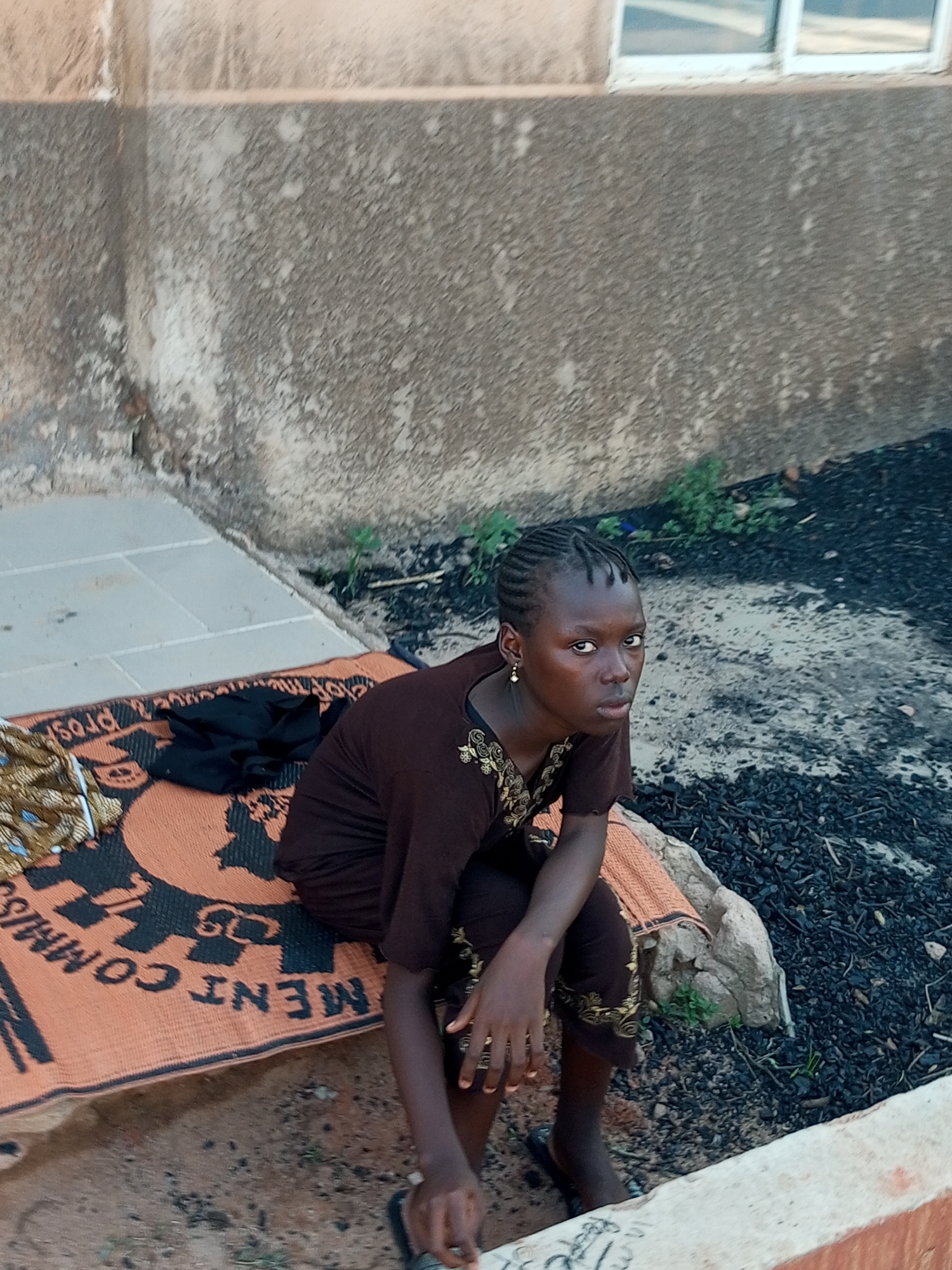 A young Black female with braided hair, wearing a brown garment with gold embroidery and a single dangly earring, sits on an orange mat with a black and white pattern. The mat is placed on a stone slab next to a textured concrete wall. Behind her, the ground is covered in small black pebbles and sparse green vegetation. To the left, a patterned fabric and a black garment are laid out on the mat. In the background, a window with white panes is visible against the upper part of the wall. The overall setting appears to be an outdoor area in Gombe, Nigeria, with elements of urban infrastructure and natural ground cover.