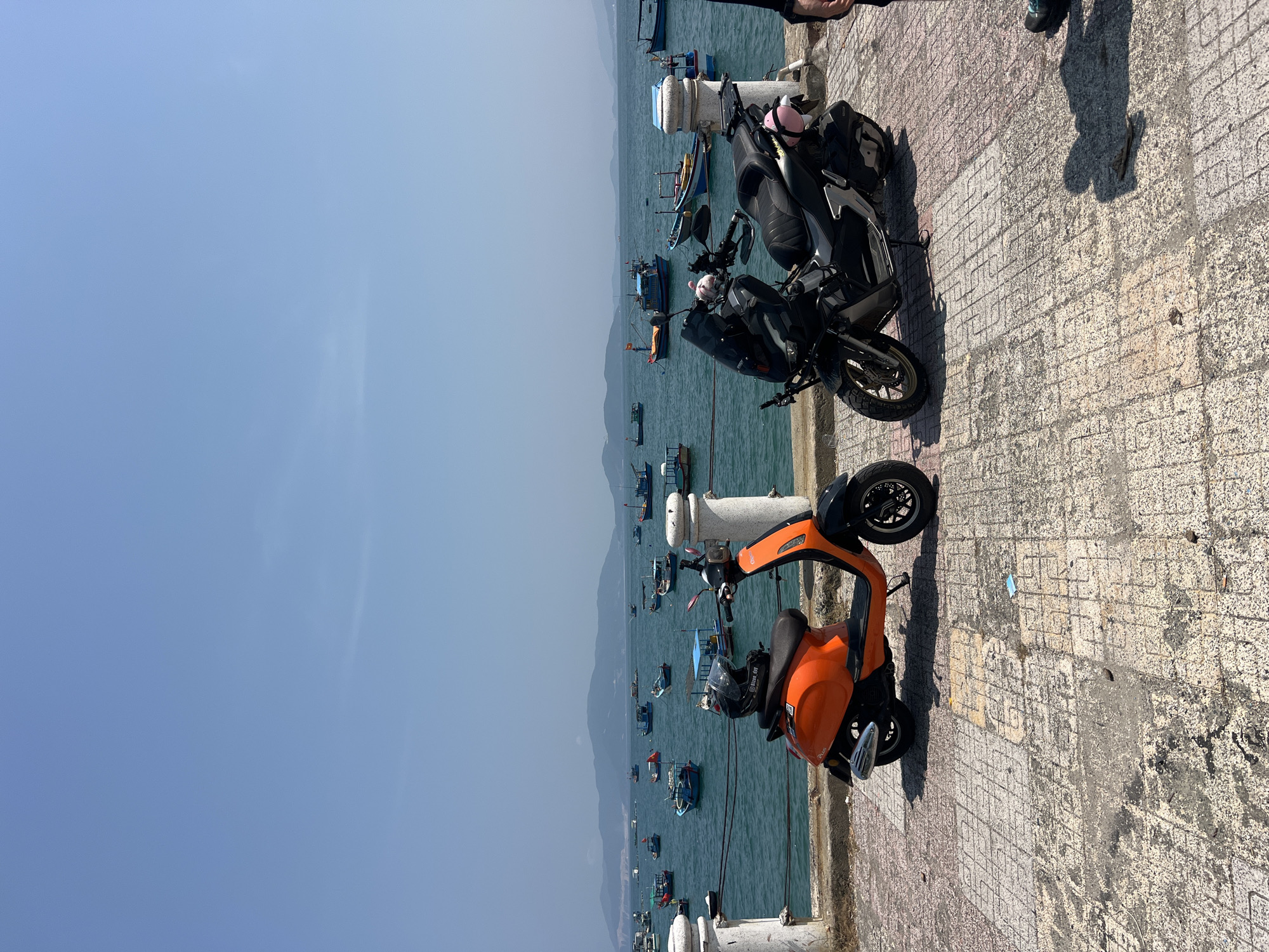 The image shows a waterfront scene in Bắc Nha Trang, Vietnam, featuring two parked scooters on a concrete walkway. An orange scooter sits closest to the viewer, while a black scooter is parked behind it. A row of white concrete bollards lines the edge of the walkway, separating it from the water. In the background, numerous fishing boats are scattered across the bay. Hazy mountains are visible in the distance, and the sky is a clear, pale blue. A section of a person's lower legs and feet is visible in the upper right corner of the image. The concrete walkway shows signs of wear and discoloration.