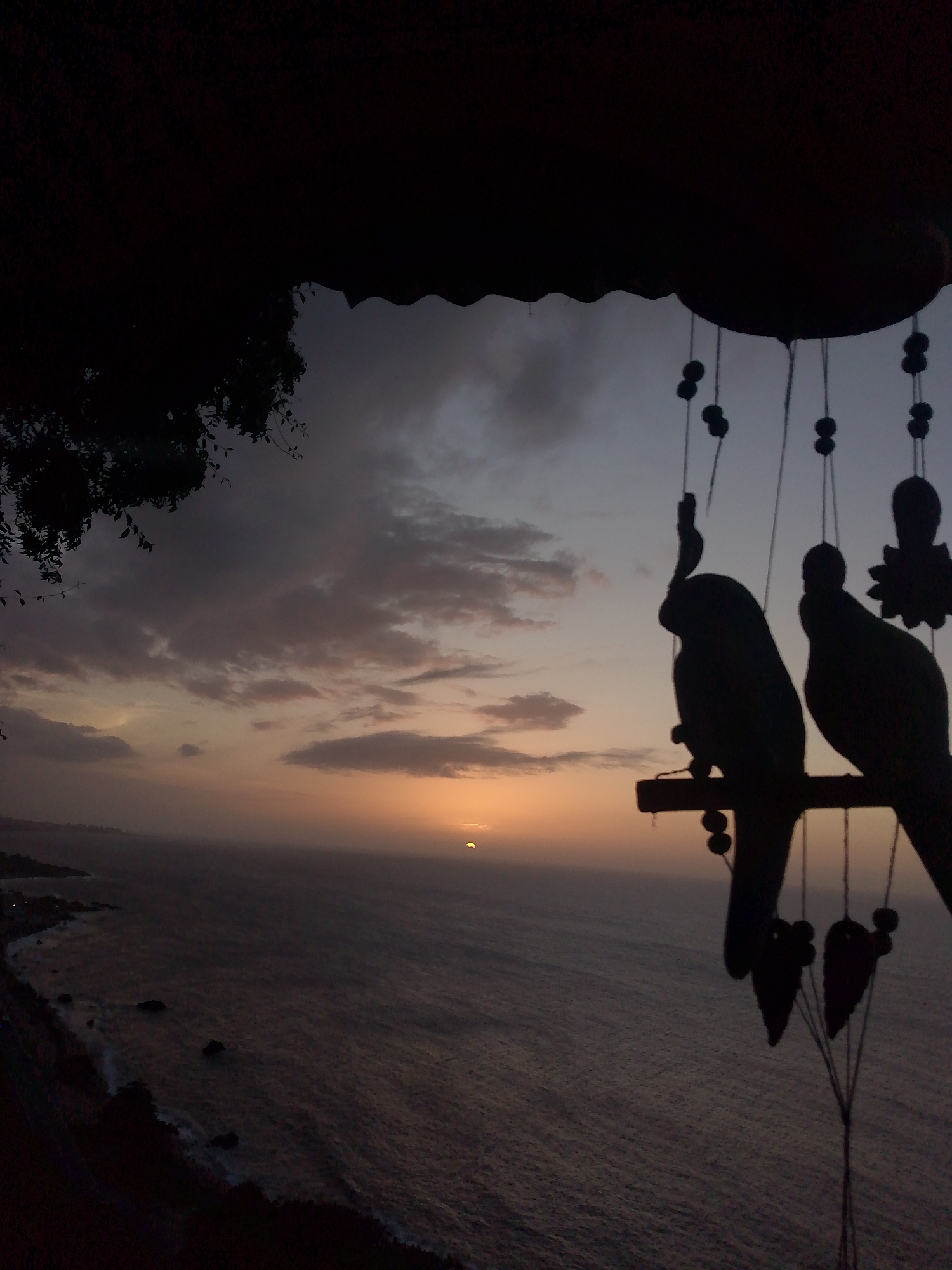 The image depicts a silhouetted view of the ocean at sunset, viewed from an elevated position, possibly a balcony or cliffside, in El Tigrillo, Venezuela. The foreground is dominated by the dark silhouettes of what appear to be decorative hanging objects, including two bird-shaped figures perched on a horizontal bar, along with other dangling shapes. The sun is low on the horizon, partially obscured by clouds, casting a warm orange glow across the sky and the vast expanse of the ocean. The coastline is visible in the mid-ground, characterized by dark landmasses and the white crests of waves breaking against rocks. The overall impression is one of tranquility and natural beauty as day transitions into evening.