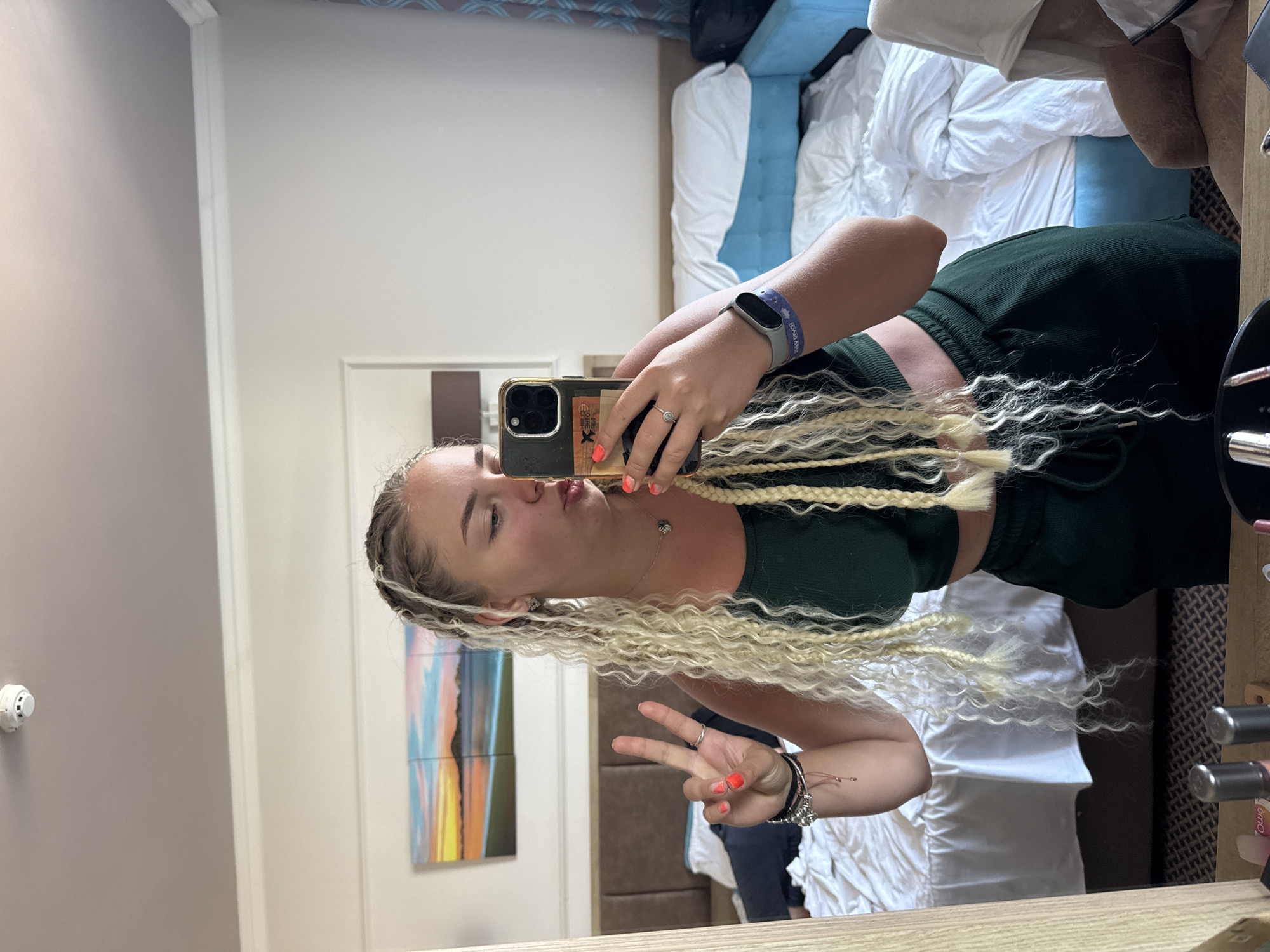 A young Caucasian woman with blonde braided hair is taking a mirror selfie in what appears to be a hotel room. She is wearing a dark green crop top and matching sweatpants. Her left hand is held up in a peace sign, and her right hand is holding a smartphone, with which she is taking the photo. She is wearing several bracelets on her left wrist and a ring on her right hand. A fitness tracker is visible on her right wrist. In the background, a bed with white linens is visible, along with a headboard and a piece of art on the wall depicting a sunset over a body of water and land. The room appears to be tidy, with a chair to the right of the bed. The lighting suggests it is daytime. The scene does not contain any elements that would specifically place it in the city of Nesebar, Bulgaria.