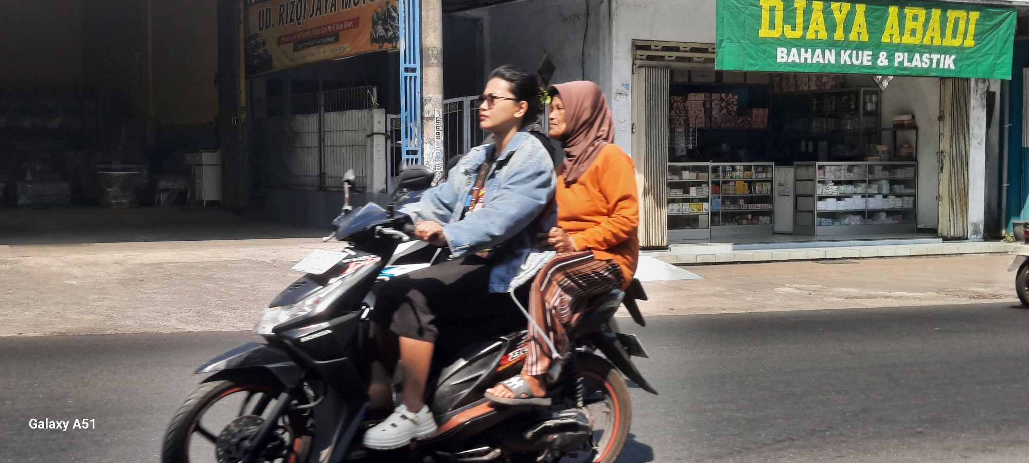 The image captures a street scene in Selopuro, Indonesia, focusing on two women riding a black motorcycle. The woman in front, wearing glasses and a denim jacket, is driving. The woman behind her, wearing an orange long-sleeved shirt and a brown headscarf, is seated as a passenger. Both women are looking to their right. The motorcycle is in motion on an asphalt road.
Behind the motorcycle, a storefront is visible with a green banner at the top that reads "DJAYA ABADI BAHAN KUE & PLASTIK". The store appears to be a shop selling baking ingredients and plastic goods, with glass display cases filled with various products. To the left of this store, another building is partially visible with a banner that reads "UD. RIZQI JAYA MOTOR". This suggests a motorcycle dealership or repair shop. The buildings line a paved street, and the overall environment appears to be a commercial area in a town or city. The lighting suggests it is daytime, with sunlight casting shadows. The lower left corner of the image displays "Galaxy A51", indicating the device used to capture the photo.