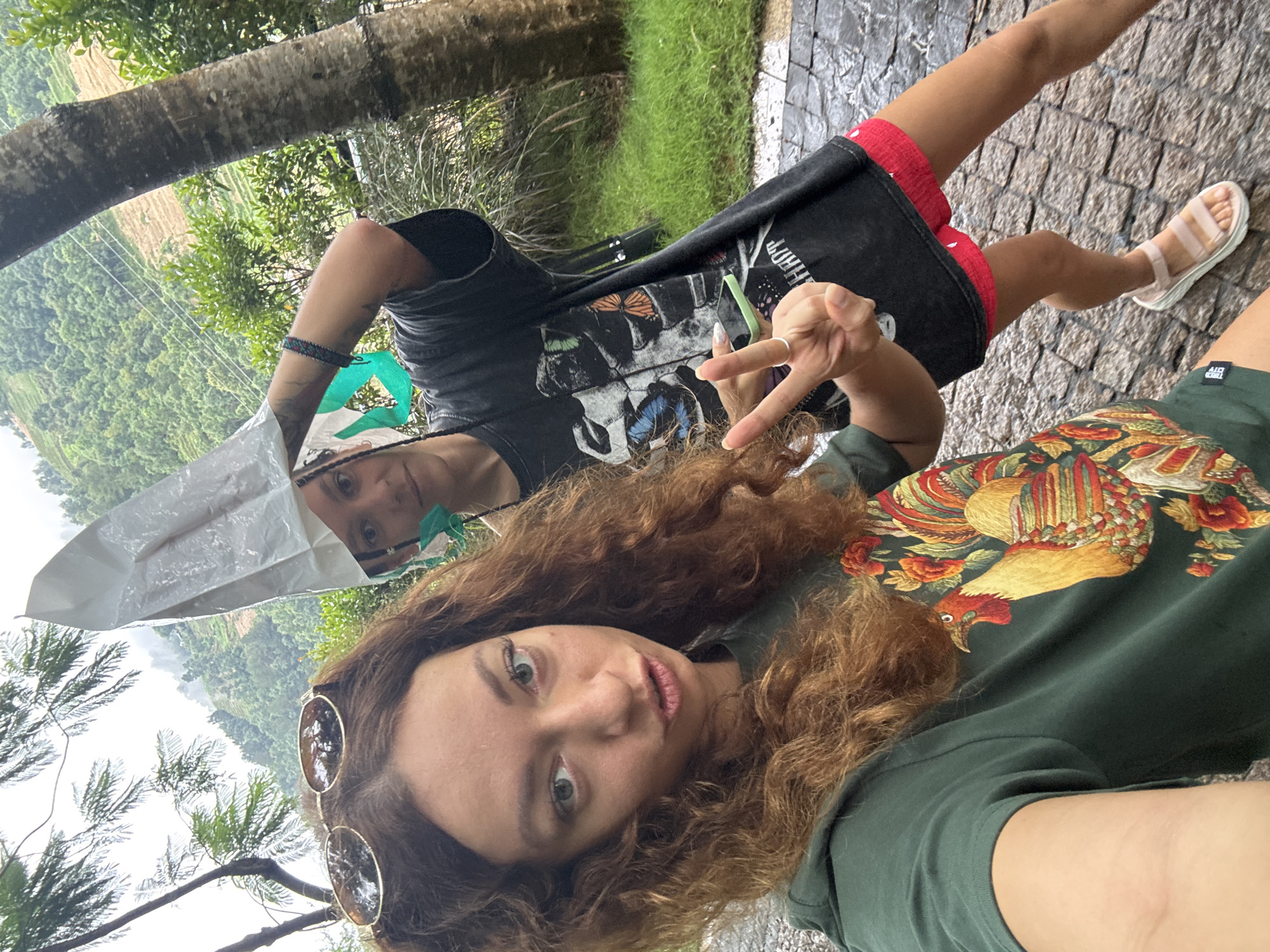 An outdoor selfie features two individuals amidst a lush, mountainous landscape, consistent with locations like Púng Luông, Vietnam. The image is rotated 90 degrees, showing the subjects in an upward-looking pose.
In the foreground, a person with long, curly, reddish-brown hair and green eyes has sunglasses perched on her head. She wears a dark green t-shirt adorned with a colorful, intricate design resembling a rooster or phoenix. A small tag on her sleeve reads "TRIBED CITY." She looks towards the camera with a neutral expression.
Behind her, another individual wears a black t-shirt featuring a prominent graphic of butterflies and a skull, with the word "BUTTERFLIES" partially visible. She sports red shorts and light pink sandals. One hand is raised, forming a peace sign, and a patterned bracelet is on her wrist. A white plastic sheet is held over her head, suggesting protection from rain or sun.
The background showcases dense green foliage and sturdy tree trunks. Beyond, rolling hills or mountains are patterned with what appear to be terraced fields. The sky is bright but possibly overcast. The ground is a paved surface of dark, textured stones or cobblestones. The presence of the plastic sheet over one person's head hints at potentially rainy or humid weather.