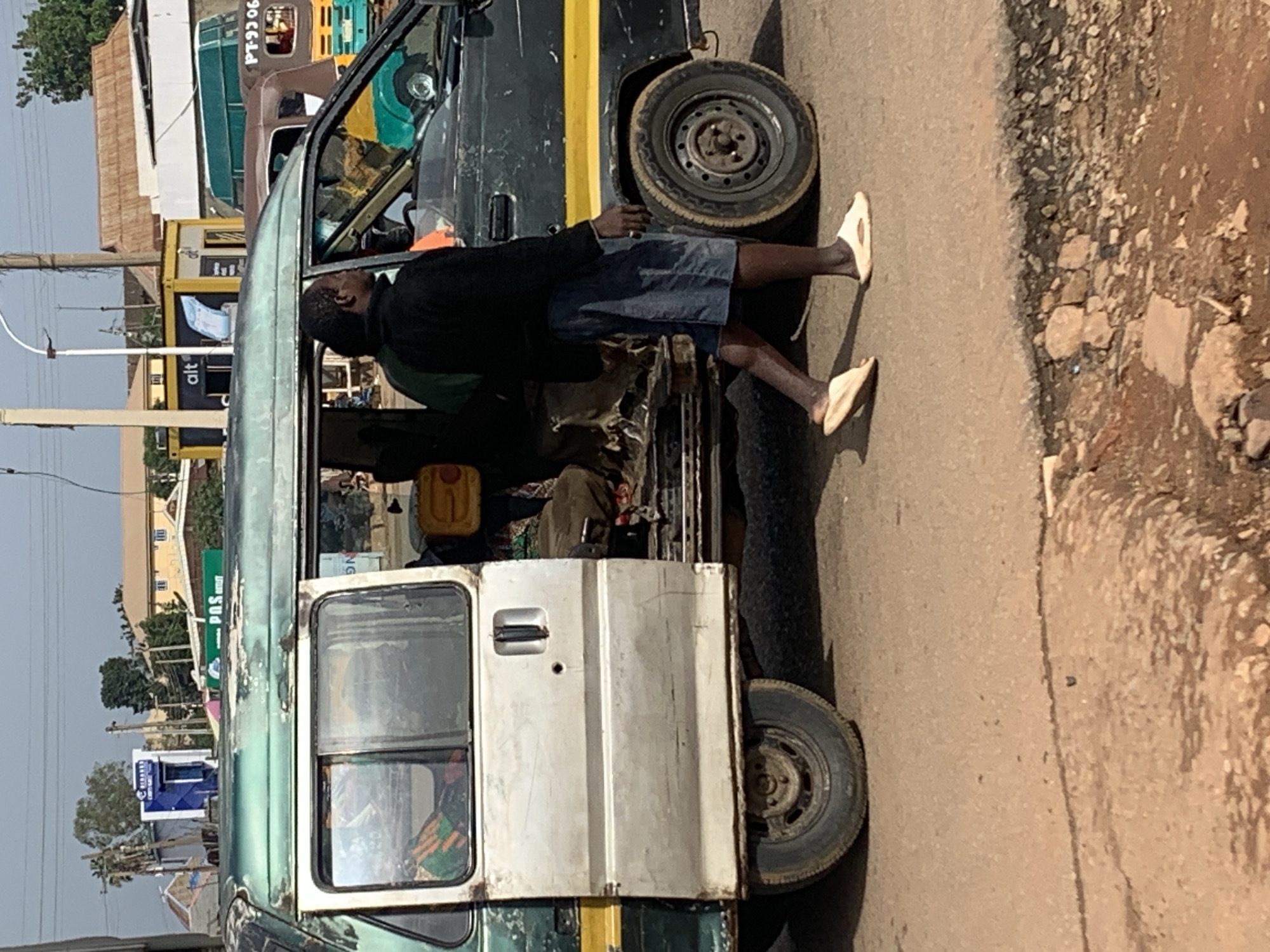 A man wearing a black jacket and denim shorts is walking next to a weathered, multi-colored van with its door open, parked on a dirt road in Gura Top, Nigeria. The van, which appears to be a form of public transportation, has a green, silver, and black exterior with a yellow stripe. Inside the van, glimpses of passengers and their belongings, including a yellow jerrycan, are visible. In the background, various buildings and structures suggest an urban environment. The road surface is uneven and dusty, with some loose stones and debris. The sky is overcast.
