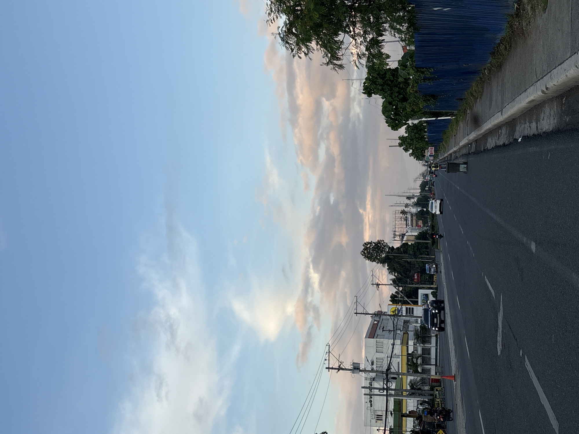 The image captures a street scene in Davao City, Philippines, likely during late afternoon or early morning, given the soft lighting and pastel colors in the sky. The sky is a dominant feature, with a light blue canvas interspersed with wispy clouds tinged with pink and orange hues.
The foreground shows a paved, multi-lane road with visible lane markings. Several vehicles are present, including a dark-colored pickup truck in the middle lane, a white sedan further down the road, and a motorcycle rider on the left side. On the right side of the road, a sidewalk is bordered by a blue corrugated metal fence. Lush green trees line the area beyond the fence, and some smaller trees are visible on the left side of the road as well.
On the left side of the street, buildings are visible, appearing to be commercial establishments, with signage and awnings. Utility poles with numerous wires are a prominent part of the infrastructure, stretching along the side of the road. There are also streetlights visible.
In the middle ground, a person is seen pushing a trash cart along the sidewalk on the right side of the road. The overall impression is of an active urban environment, likely a major thoroughfare, with everyday life unfolding under a scenic sky.