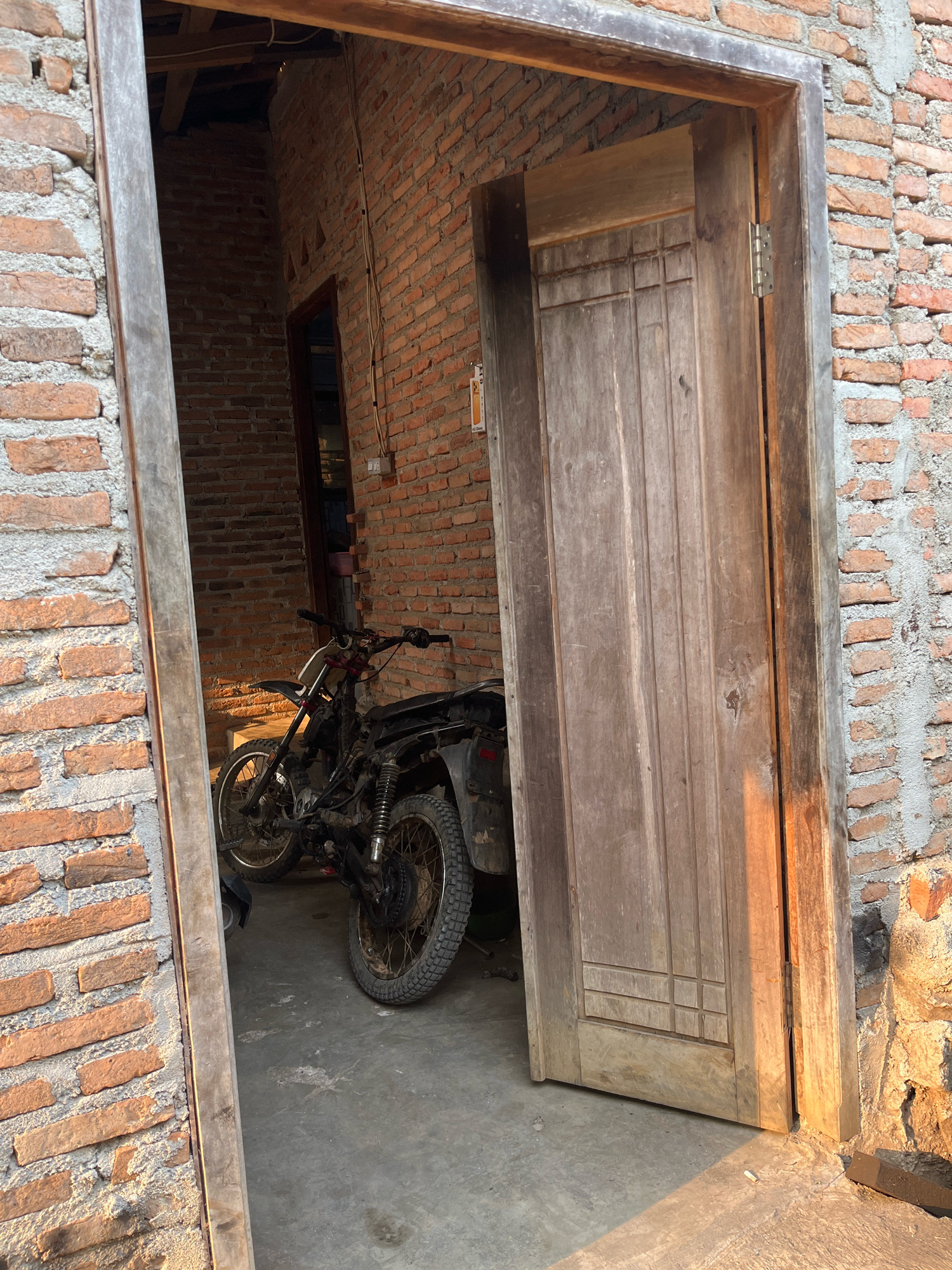A weathered wooden door stands open, revealing a motorcycle parked inside a brick structure in Kebumen, Indonesia. The brick walls are visible on either side of the doorway, with rough, unfinished mortar. The motorcycle appears to be a dirt bike or enduro model, with knobby tires and a relatively upright riding position. It is dark in color and shows signs of wear. The interior space appears to be a simple, open area with a concrete floor. Sunlight casts warm shadows on the door and the surrounding brickwork, suggesting it is late afternoon or early morning. The scene evokes a sense of rustic simplicity and perhaps a utilitarian purpose for the space.
