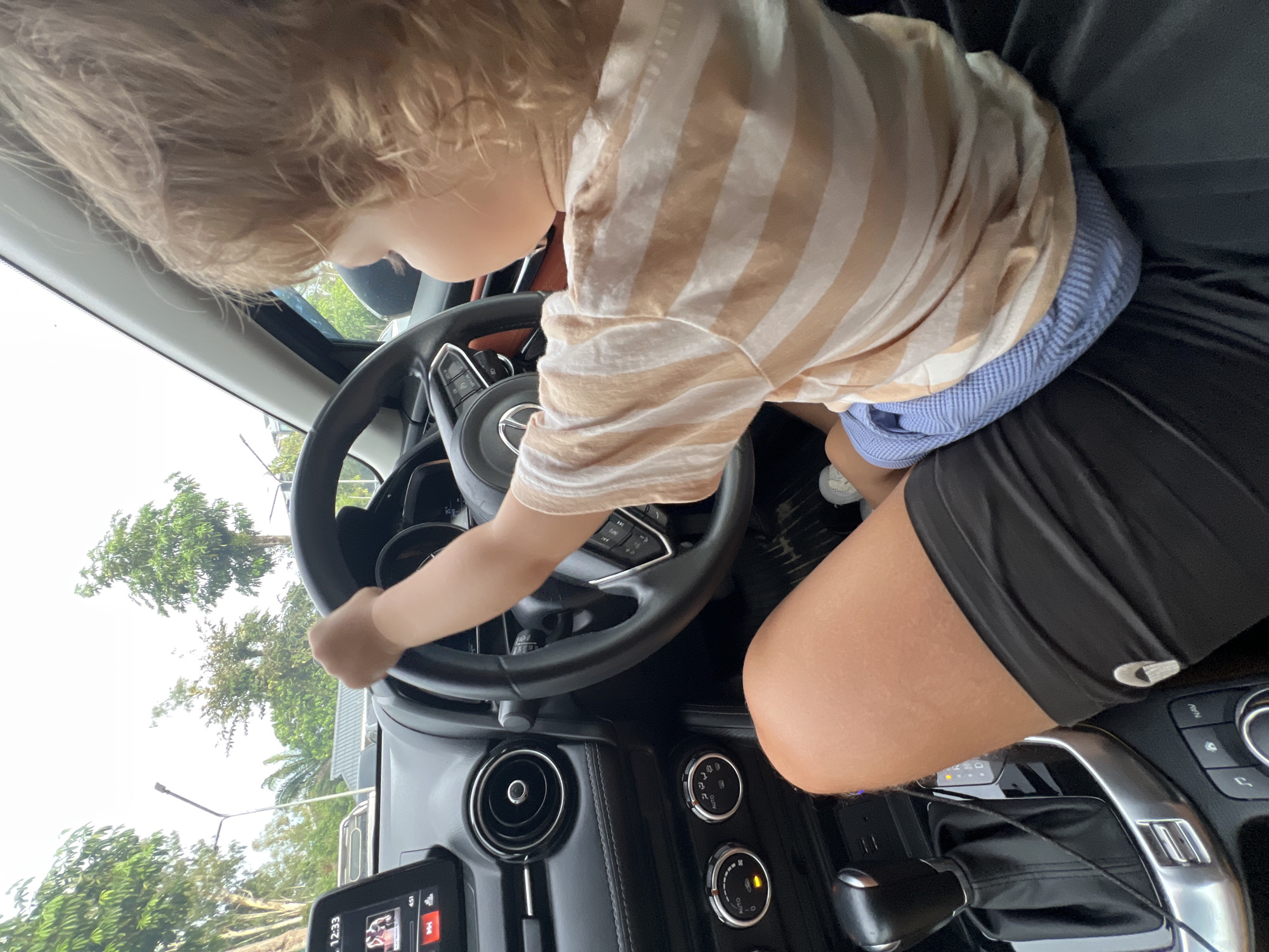 The image shows a toddler sitting on an adult's lap in the driver's seat of a car. The toddler is wearing a striped t-shirt and blue shorts and is holding the steering wheel. The adult is wearing black shorts. The car's interior is visible, including the dashboard, steering wheel, gear shift, and infotainment screen. The background shows trees and sky, indicating the car is outdoors, possibly in a parking lot or on a street. The overall scene depicts a playful interaction between an adult and a child.
