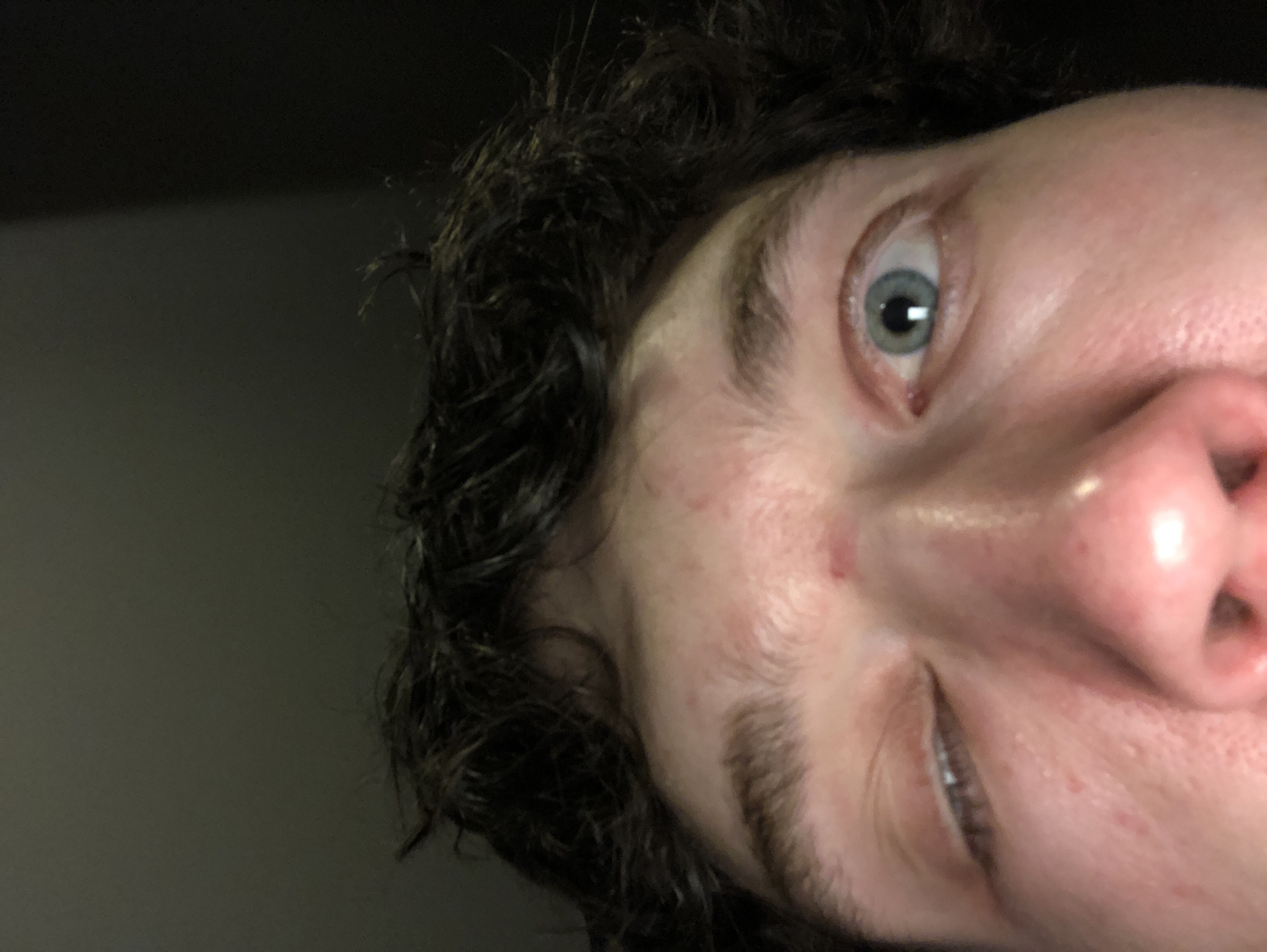 A close-up image of a person's face, angled from below. The person has dark, curly hair, fair skin, and light-colored eyes. The person's nose and right eye are prominently featured. The background is dark and out of focus. The location is Unknown, Unknown.