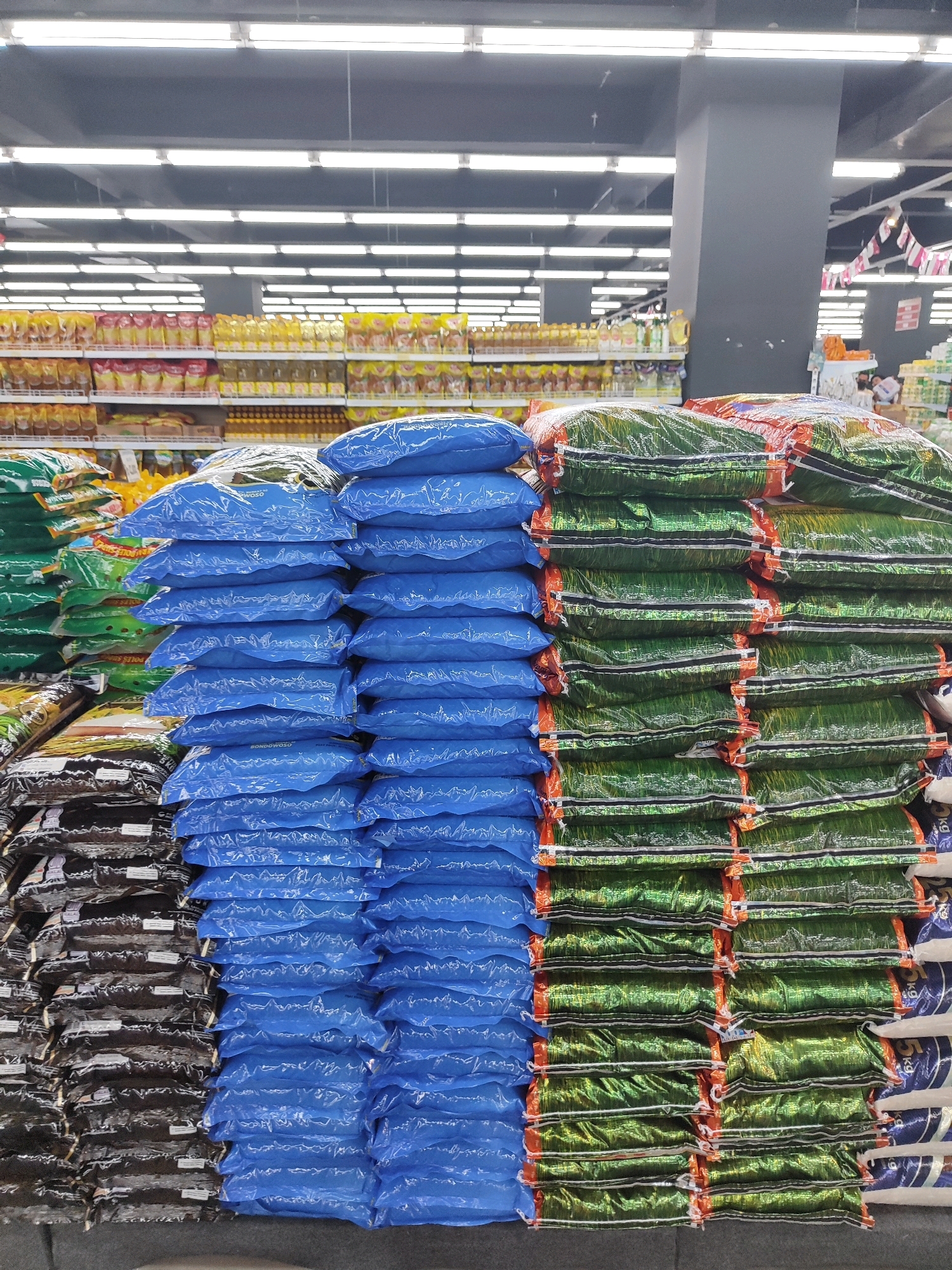 The image shows a retail store in Sukowiryo, Indonesia, with stacks of packaged goods. In the foreground, there are large, blue plastic bags stacked high, some of which are labeled with text that appears to be in Indonesian. To the right of the blue bags are stacks of green and orange plastic bags, also containing packaged goods. Further to the left, there are stacks of dark-colored bags. In the background, shelves are visible, stocked with various food products, including bottles of cooking oil and packets of seasoning. The store appears to be well-lit with fluorescent lighting fixtures on the ceiling. The overall scene depicts a commercial environment within a grocery or supermarket setting.