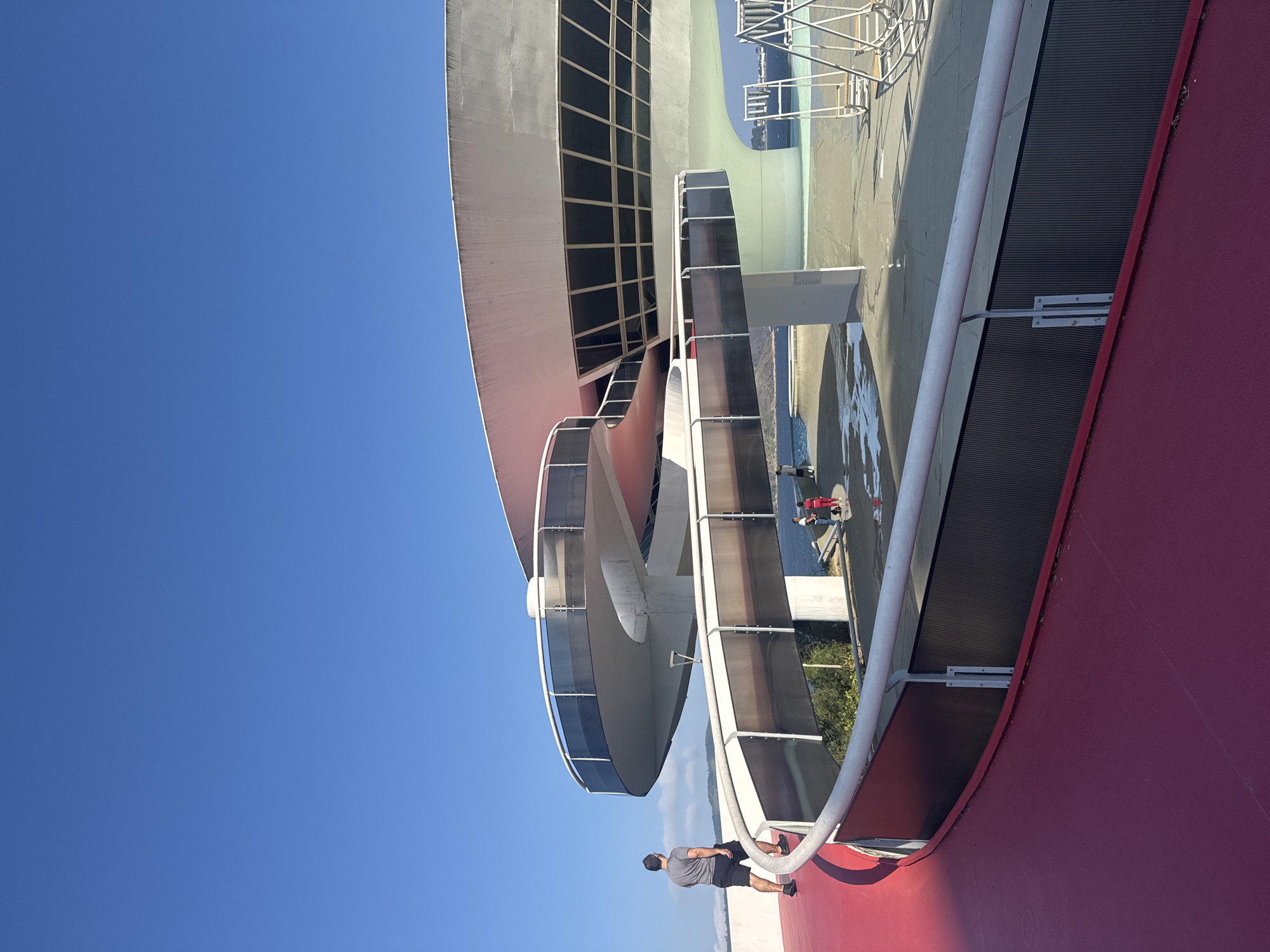 This is an outdoor, ground-level, eye-level shot of the Museu de Arte Contemporânea de Niterói (MAC), designed by Oscar Niemeyer, located in Niterói, Brazil. The museum is characterized by its circular, saucer-like shape. A winding red walkway with a white railing curves into the foreground from the left, leading towards the museum. A man in a grey t-shirt and black shorts walks away from the viewer on this red walkway. Further back, people can be seen standing near the base of the museum. The museum building itself is white with a circular structure and a facade of dark, rectangular windows. A body of water is visible behind the museum. The sky is a clear, bright blue.