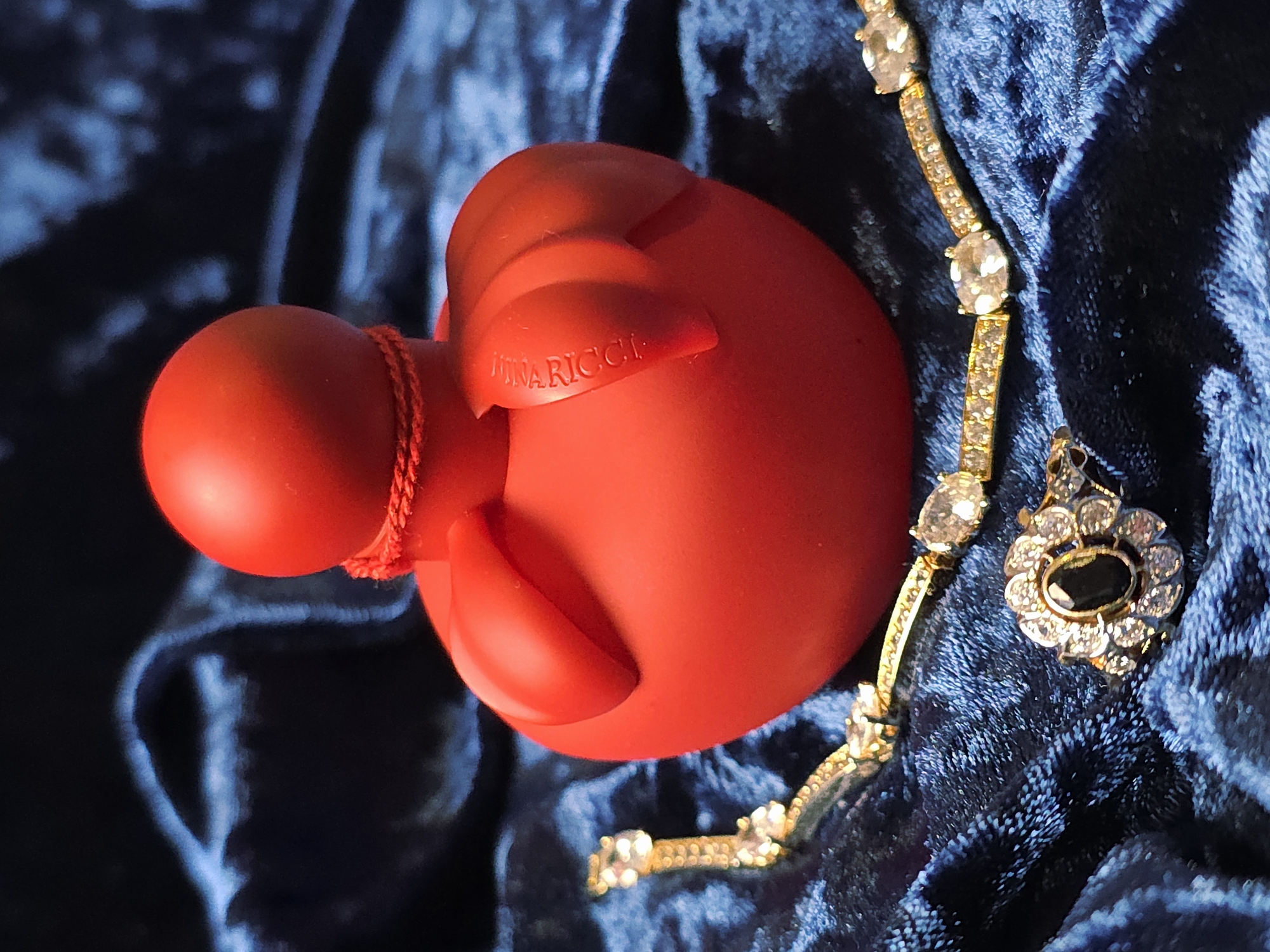 A red Nina Ricci L'Extase perfume bottle sits on a dark blue velvet surface next to a gold bracelet with clear stones and a pendant featuring a dark stone surrounded by clear stones.