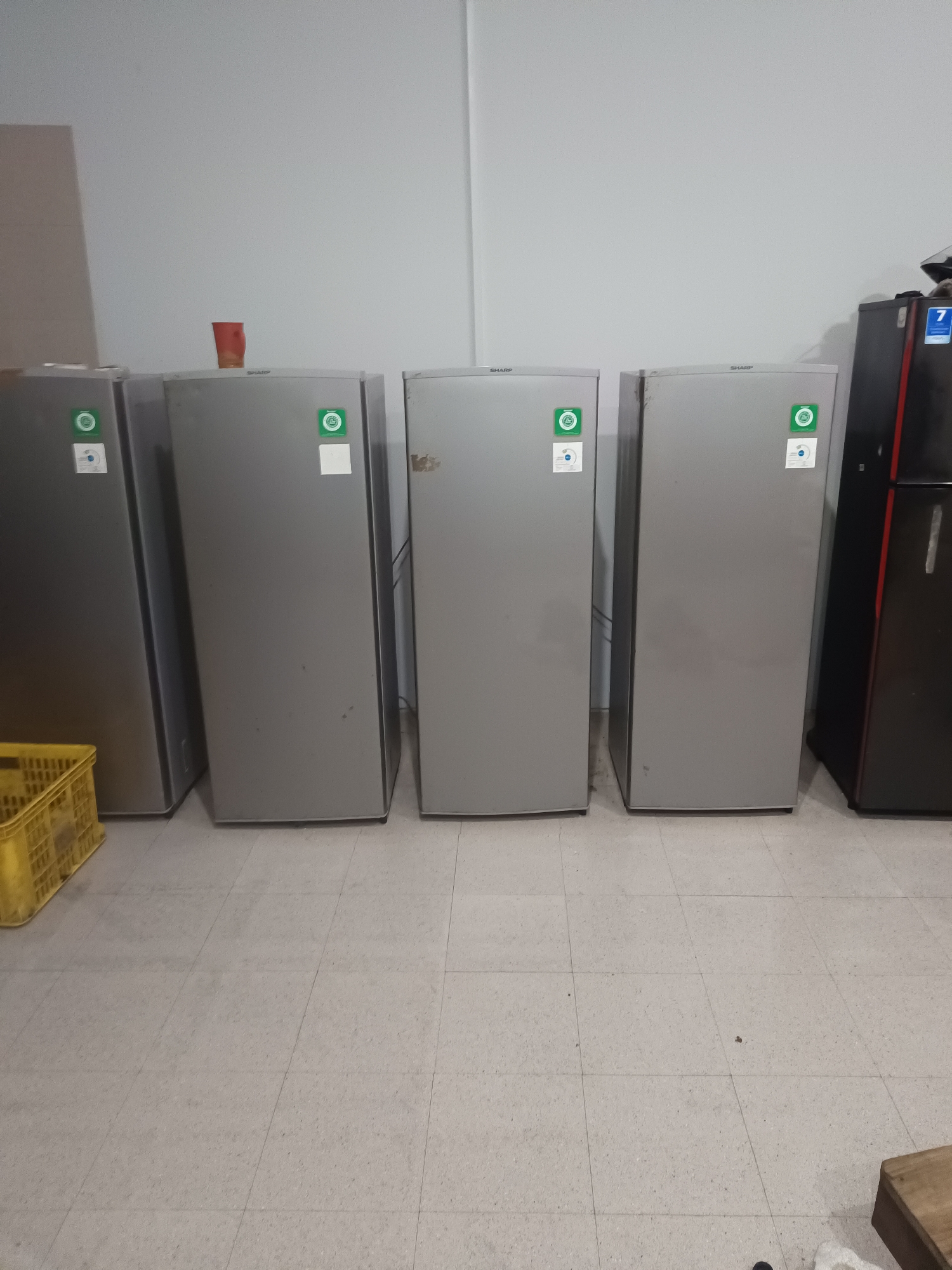 A row of at least five tall, narrow, silver-colored refrigerators are standing against a plain, light-colored wall. The refrigerators are evenly spaced and appear to be new or in good condition, with green and white circular stickers on the upper portion of each door. The floor is tiled with light gray square tiles. In the foreground, to the left, a yellow plastic basket is partially visible. To the far right, a dark-colored refrigerator with a red accent is also present. A single red plastic cup is placed on top of one of the silver refrigerators. The scene suggests a storage area or a showroom for appliances. The image does not contain any visible people or evidence of a specific location within North Jakarta, Indonesia.