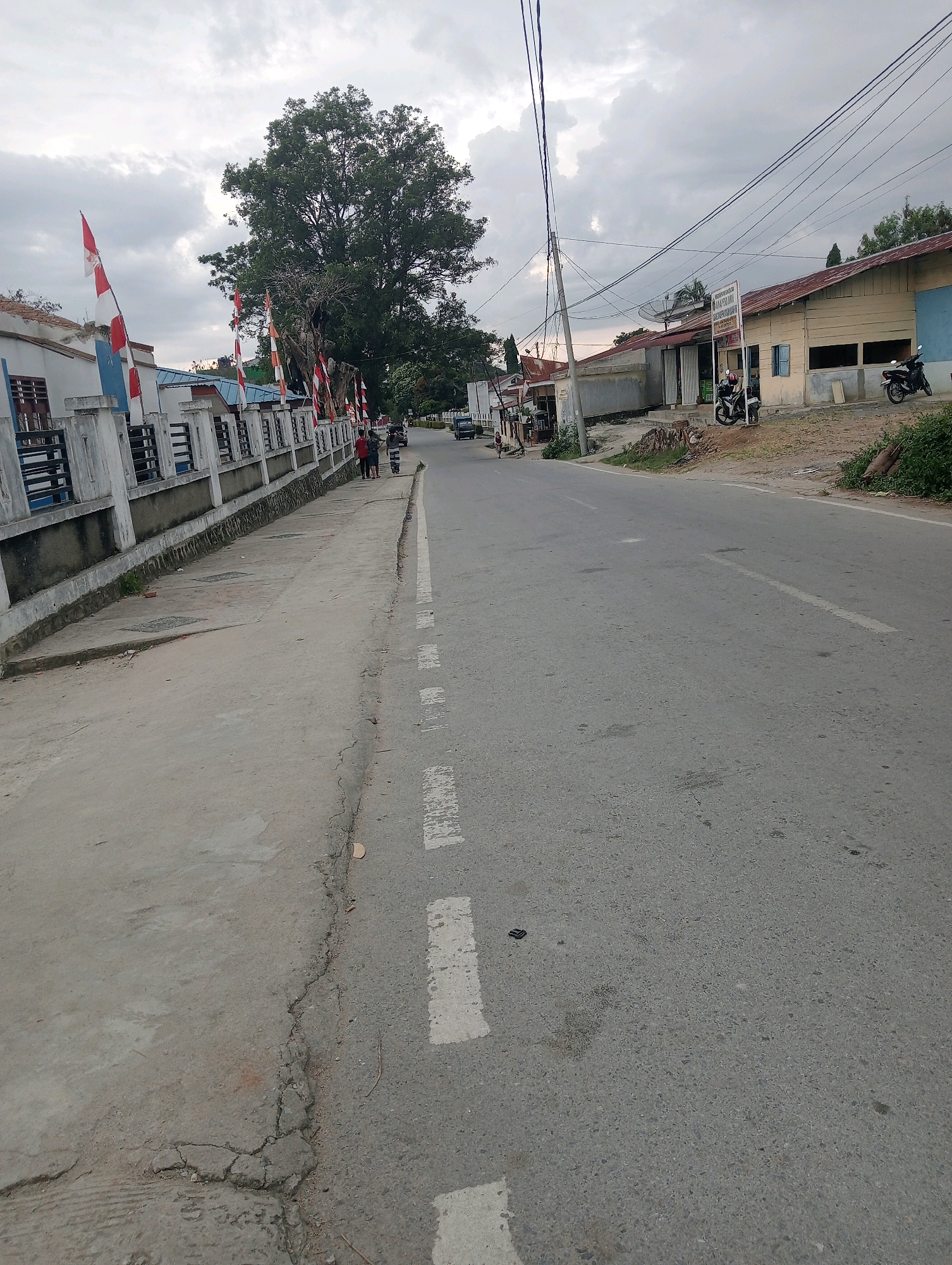 The image depicts a street scene in Hinalangbagasan, Indonesia, likely during overcast weather. On the left side of the road, a white concrete fence with black bars lines a sidewalk. Several Indonesian flags are displayed along the fence. A large tree with dense foliage stands behind the fence. The road itself is paved with asphalt and has white dashed lines marking lanes. On the right side of the road, several buildings with reddish-brown roofs are visible. One building has a sign that appears to be in Indonesian. A satellite dish is also present on one of the roofs. Parked motorcycles are visible in front of the buildings, and a few people are walking on the sidewalk. The overall impression is that of a rural or semi-urban setting.