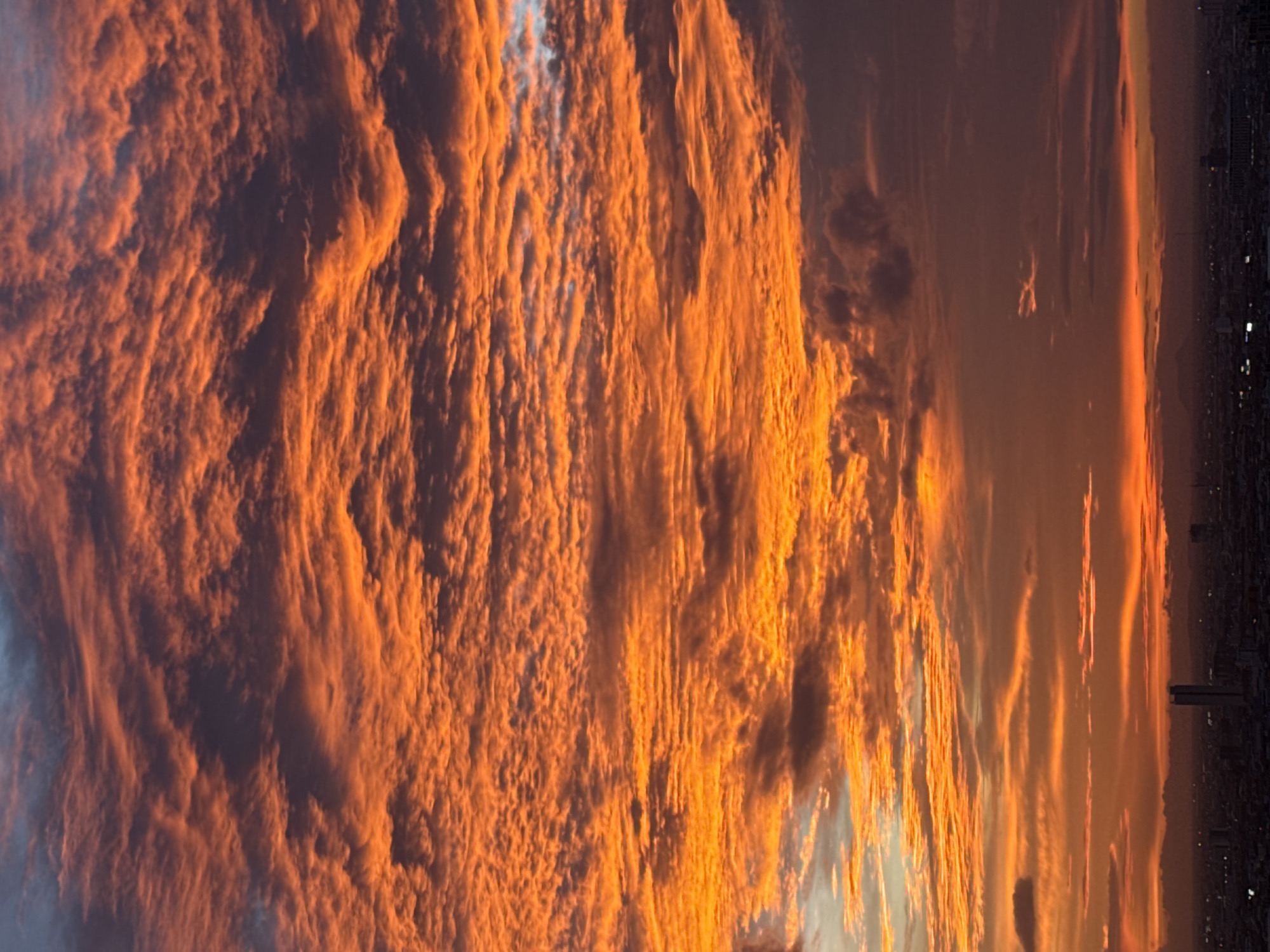 The image depicts a dramatic sunset over the city of Khan Mean Chey, Cambodia. The sky is filled with vibrant orange and red clouds, with some darker, purplish clouds interspersed. The clouds are textured and appear to be in multiple layers, creating a sense of depth and movement. Below the sky, a silhouetted cityscape is visible. Buildings, including a tall, slender skyscraper, are discernible against the twilight sky. The horizon line is marked by a range of dark hills or mountains in the distance. The overall scene conveys a sense of natural beauty and urban landscape at dusk.