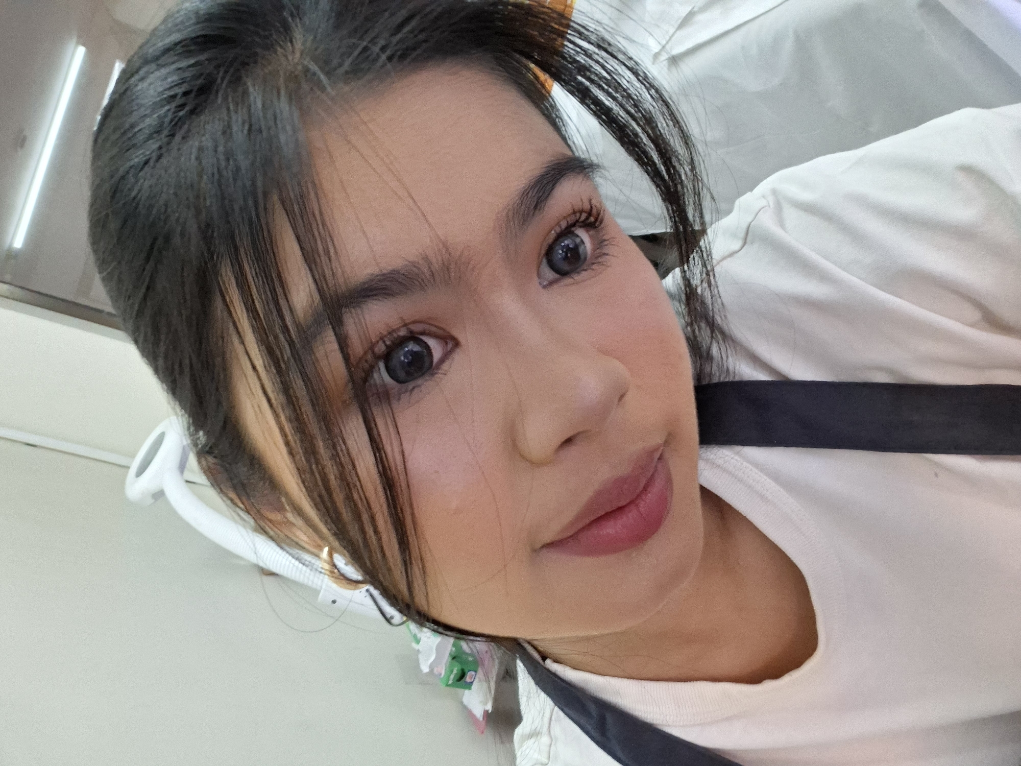 A close-up, selfie-style, portrait shows the face of a young woman with dark hair pulled back, some strands falling around her face. She is wearing a white t-shirt and a dark apron strap is visible across her chest. Her eyes are dark and wide, and she has bright pink lipstick on. She is looking towards the camera with a slight smile. In the background, a white light fixture with a flexible arm is visible, attached to a light-colored wall. There are also some blurry objects in the background, including a green box and what appears to be part of a white garment. The overall environment suggests an indoor setting, possibly a salon or a similar service establishment. The image was taken in Kuta, Indonesia.