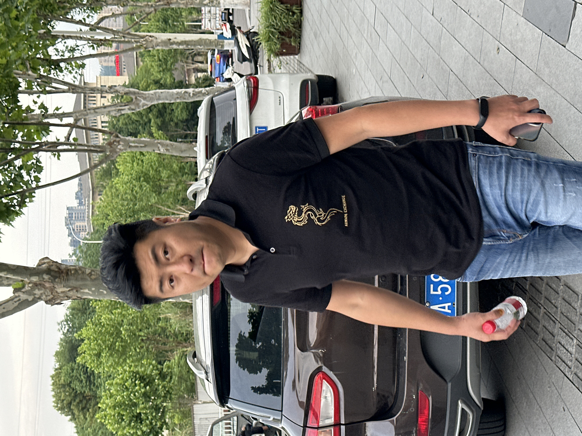 An East Asian man stands next to a dark-colored SUV on a paved sidewalk, looking directly at the camera. He is wearing a black short-sleeved polo shirt with a gold dragon motif and "ARMANI EXCHANGE" text on the left chest, paired with blue jeans. A black wristband is on his left wrist, and he holds a smartphone in his left hand. In his right hand, he holds a clear plastic bottle with a red cap.
The scene is an urban outdoor setting in what appears to be Jiangjin, Chongqing, China, during the daytime on an overcast or mildly bright day. The man is positioned in front of the rear of a dark SUV, which has a blue license plate partially visible, reading "重庆 A.59XX", indicating a vehicle from Chongqing. Behind this SUV, a white SUV is parked, and further back, an electric scooter is visible. The street is lined with mature plane trees featuring their characteristic peeling bark and green foliage. In the background, multi-story buildings and a city skyline are visible under a light sky. A plant pot with green vegetation is also seen in the background. The ground is covered with light grey square tiles. The man's expression is neutral to slightly serious.
