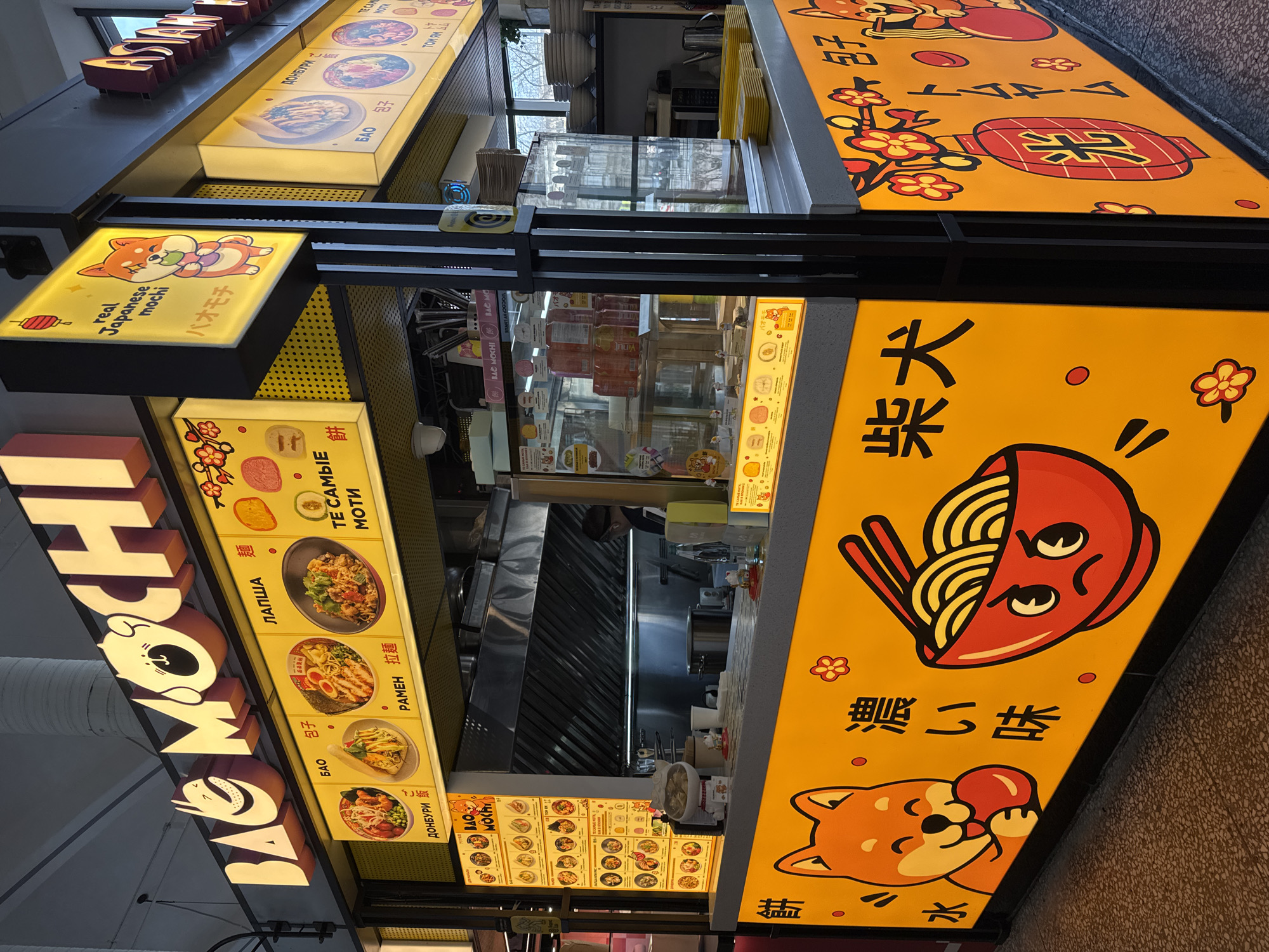 The image shows a food stall in what appears to be a shopping mall in Saint Petersburg, Russia. The stall is brightly lit with signs in both Russian and Japanese or Chinese characters. The name of the stall, "BAO MOCHI," is prominently displayed with cartoon characters. Menu items such as bao, lapsha (noodles), ramen, and donburi are advertised, with illustrations and Russian text descriptions. A sign depicts a Shiba Inu dog eating a mochi. Other signs mention "real Japanese mochi." The stall's interior is visible with kitchen equipment, utensils, and packaged food items. A price menu is posted with various dishes and prices in Russian. The location is likely inside a shopping center or food court given the lighting and infrastructure.