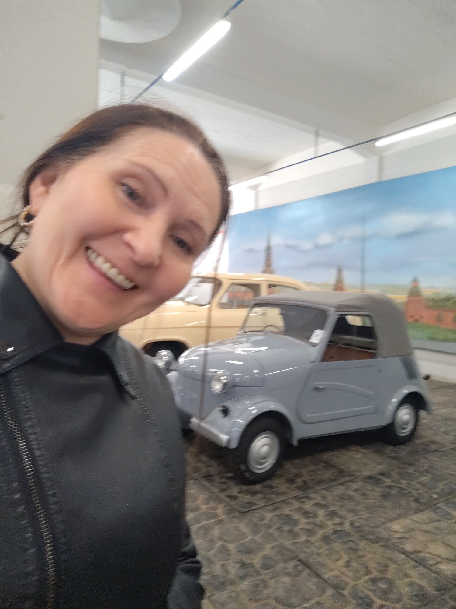 A woman is captured in a selfie in an indoor setting, likely a museum or exhibition hall. She is in the foreground, smiling broadly with her teeth showing, and appears to be happy or amused. She has dark hair, wears a black collared jacket (possibly leather), and a gold hoop earring is visible on her left ear.
In the background, two vintage cars are on display. The more prominent vehicle is a small, grey, two-door convertible with a dark fabric soft top. It has classic styling, round headlights, and silver hubcaps. Behind it, partially visible, is a larger, light beige-colored car, possibly an older sedan or station wagon.
The cars are set against a large painted mural depicting a cityscape with red-brick walls and multiple towers with spires, characteristic of the Moscow Kremlin, under a blue sky with clouds. The floor beneath the cars is dark and patterned, resembling cobblestones or a paved area. The ceiling is white with bright fluorescent tube lights, indicating an artificially lit indoor environment.