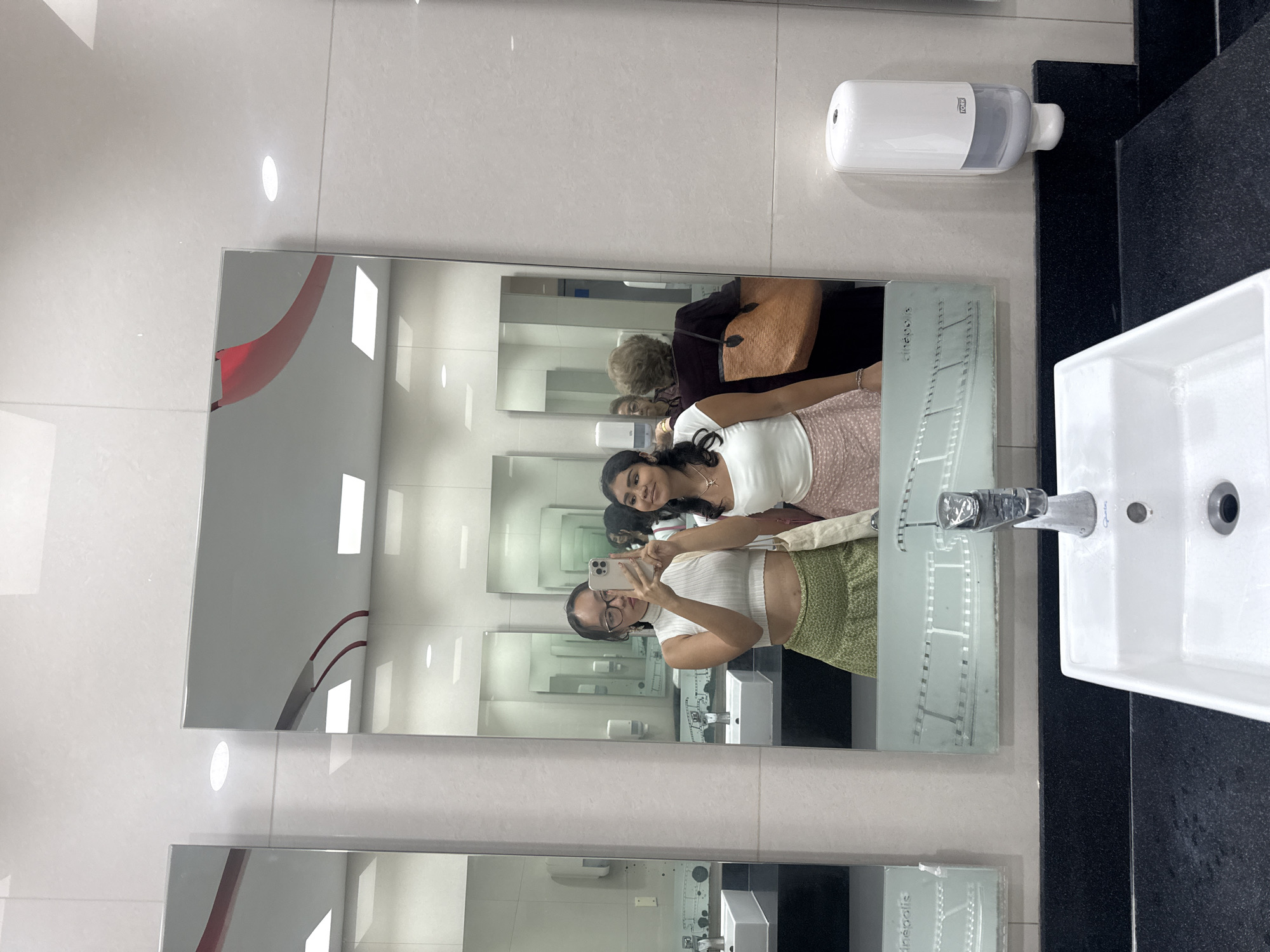 Two women are taking a selfie in front of a mirror. The reflection in the mirror indicates the location is likely a public restroom, possibly at a Cinepolis movie theater in Puebla, Mexico, as "Cinepolis" is etched on the mirror. One woman is holding a cell phone and appears to be taking the photo. The other woman is smiling and posing. An older woman with a tan purse is visible in the background, also reflected in the mirror. A white sink, faucet, and soap dispenser are present in the foreground. The walls are white tiles.