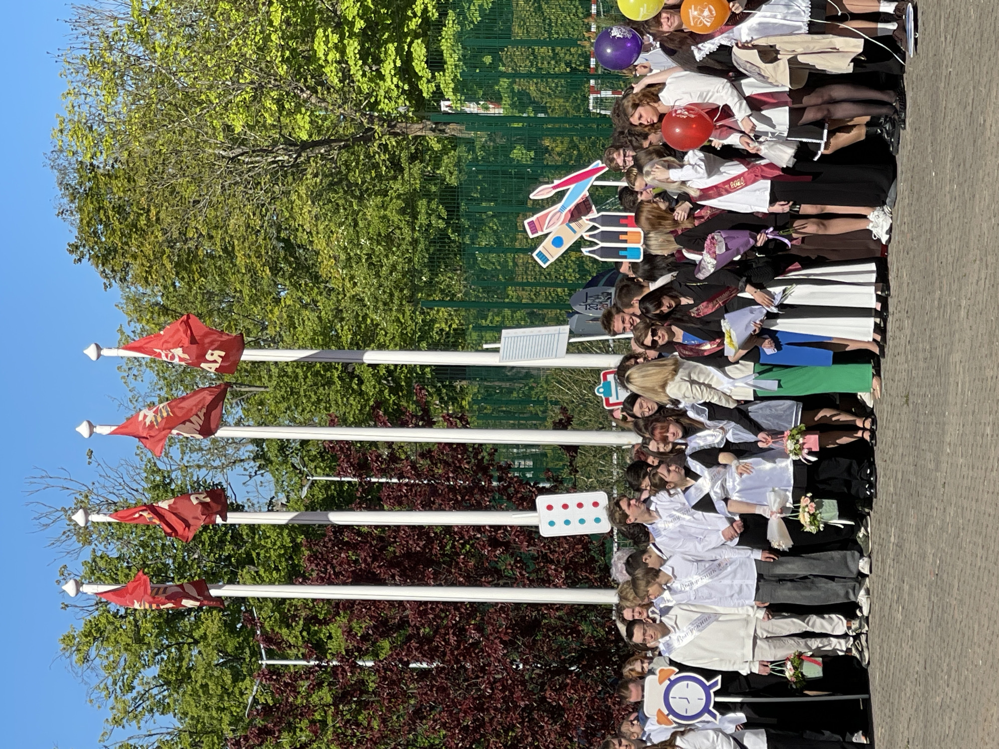 A horizontal, eye-level shot reveals a crowd of people, predominantly young adults, positioned perpendicular to the camera, standing outside in Zelenogradsk, Russia. Most people are wearing white shirts and dark pants or skirts, with some sporting sashes that read "Выпускник" and "2022" in Cyrillic, indicating graduation. Some carry bouquets of flowers, signs, and balloons. In the background are green trees, a green metal fence, and a blue sky. In the foreground, there are four flagpoles, each with a red flag bearing white Cyrillic text. The setting appears to be a public space, possibly a schoolyard, with a brick pathway visible in the lower right.