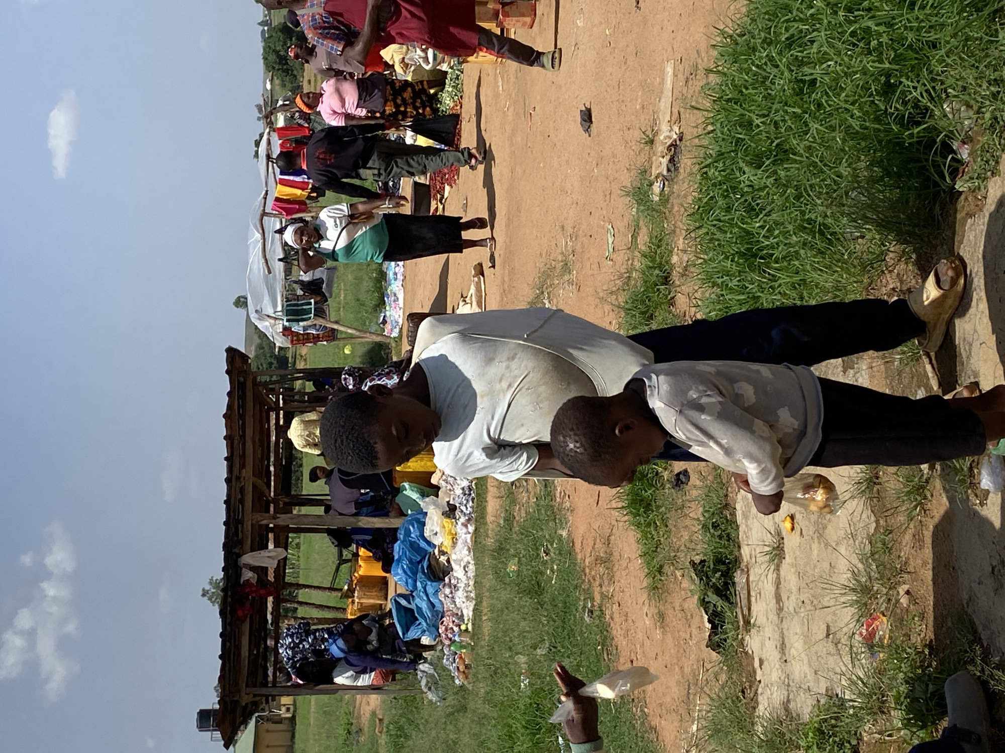 The image shows an outdoor marketplace scene in Riyom, Nigeria. Two people, possibly a parent and child, stand near the foreground. The child appears to be eating something from a clear plastic bag. In the background, other people are browsing stalls beneath a makeshift shelter. Merchandise such as clothing and blue tarps are visible. The ground is dirt and grass.