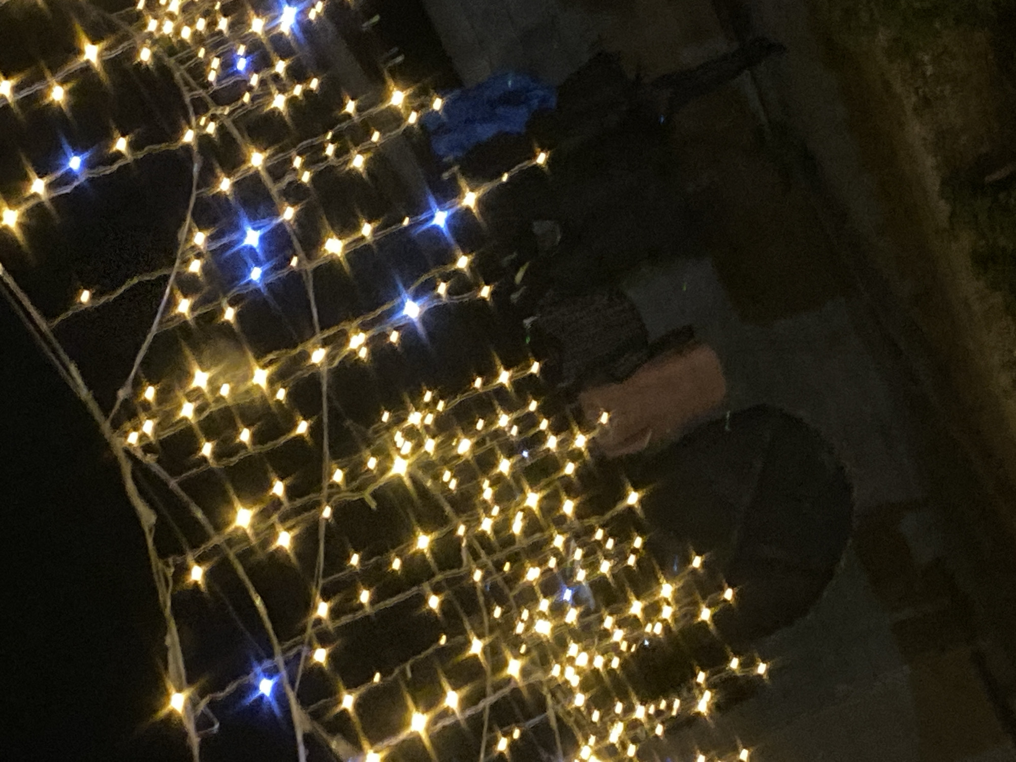 An outdoor scene at night is illuminated by numerous strings of warm yellow and some blue fairy lights, creating a bokeh effect against a dark background. The lights are draped and strung across the upper portion of the frame, casting a glow on objects and indistinct figures below. In the mid-ground, partially obscured by the lights, are several dark, silhouetted forms that appear to be people wearing dark clothing. One figure on the right is wearing a bright blue garment. To the left of center, a tan-colored object, possibly clothing, hangs amongst the lights. The ground appears to be dark pavement or dirt. The overall impression is that of a festive or decorated outdoor space at night. The location is the city of Babayurt, Russia.
