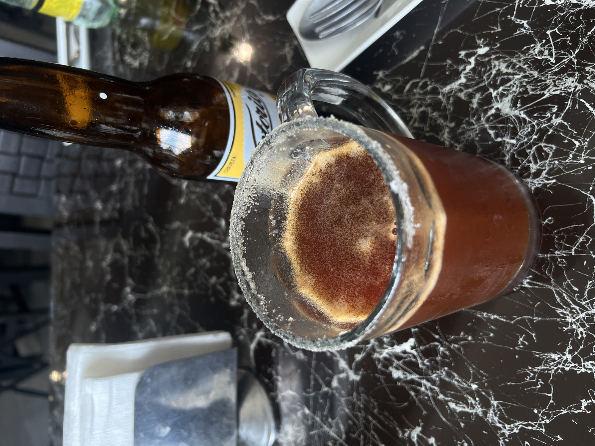 A top-down shot displays a beer bottle and a beer mug rimmed with salt on a black marble-patterned table. The beer bottle is brown with a yellow label. The mug contains a reddish-brown liquid, likely beer. Beside the beer bottle is a white container holding silverware. A stack of white napkins sits near the bottom left of the image. The location is assumed to be in Mexico City, Mexico.