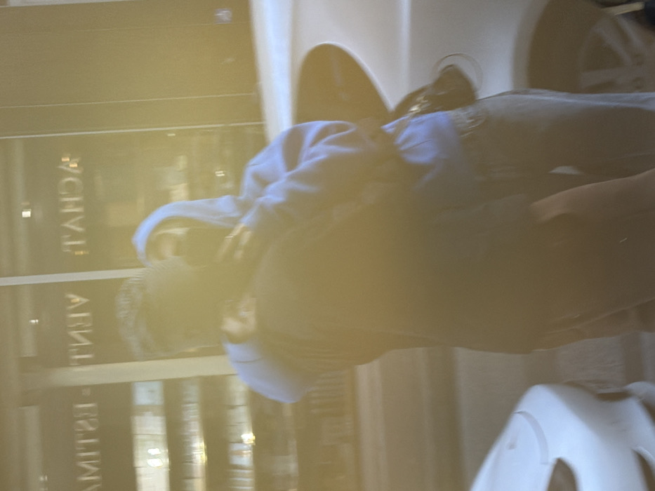 A person wearing a blue hooded sweatshirt and jeans sits in a wheelchair. The person has light hair and is facing towards the right of the frame. A store window is visible in the background, with reflective writing. A white object is near the bottom right of the image, possibly part of the wheelchair or an adjacent object. The image has a blurred, golden hue.