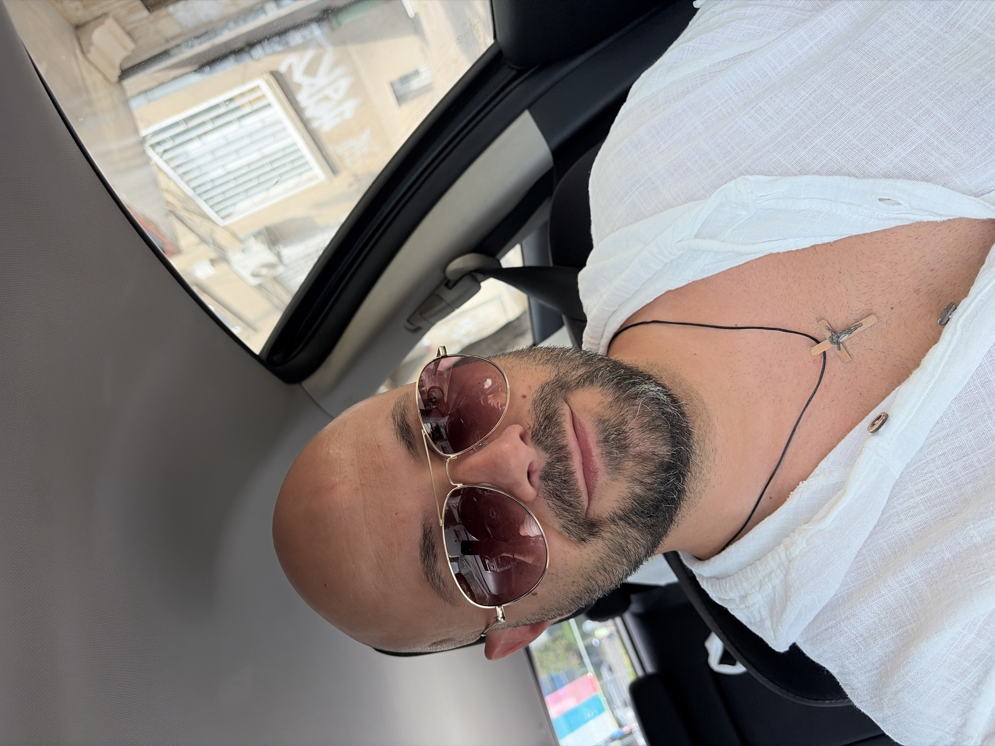 The image shows a man sitting in the driver's seat of a car, taking a selfie. He is bald, has a beard, and is wearing aviator sunglasses and a white linen shirt. He is wearing a necklace with a wooden crucifix. The car appears to be parked on a street in a city, with buildings and windows visible through the car's windows. The man's reflection is visible in the sunglasses, showing him holding the phone and what appears to be a car interior. The overall scene is mundane and personal, capturing a moment in the man's day. The presence of graffiti on the building visible through the window suggests an urban environment, possibly in an older part of the city.