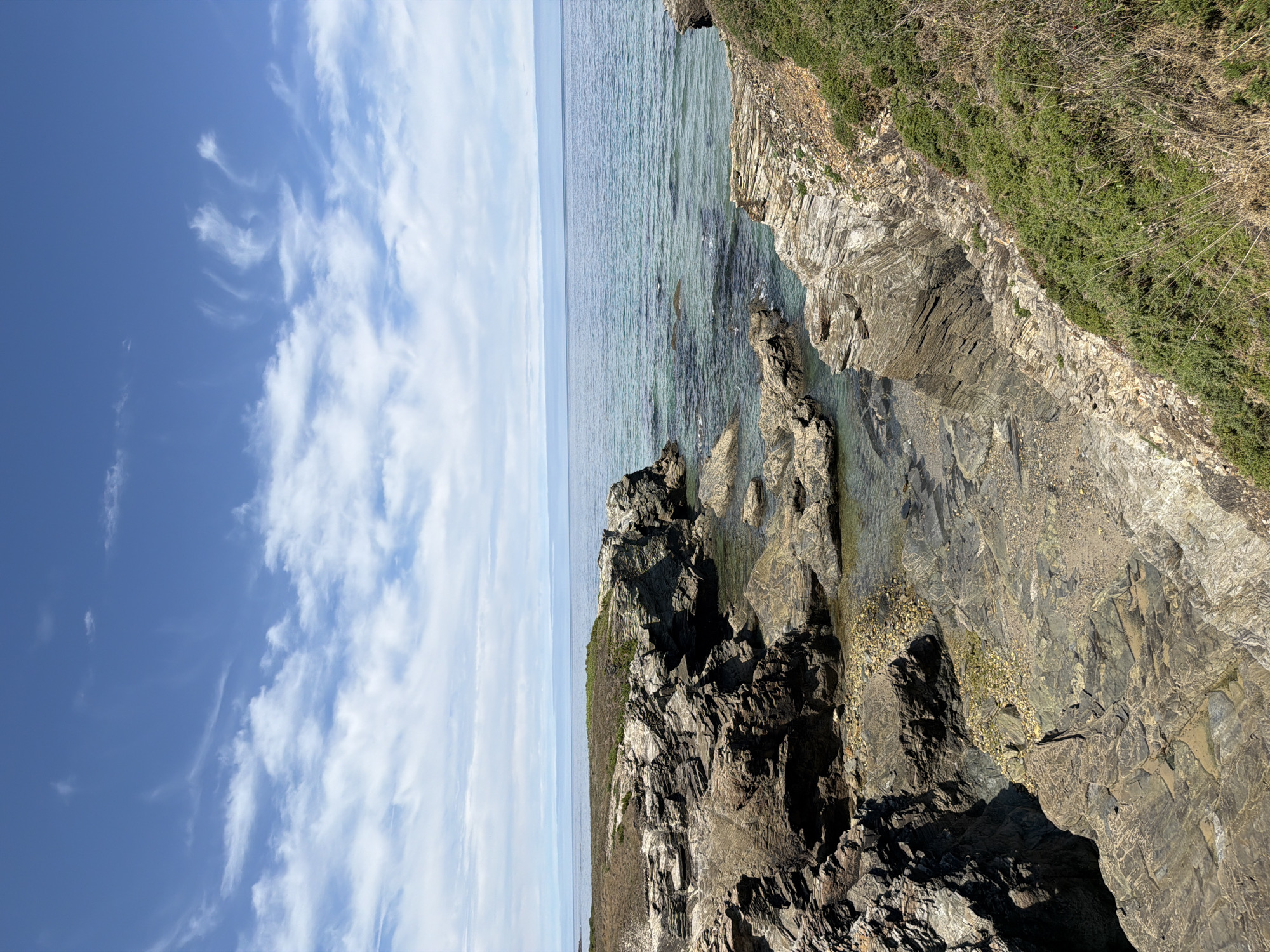 The image depicts a rocky coastline with the ocean under a blue sky with scattered clouds. The foreground shows layers of gray and brown rock formations, some of which are covered with sparse green vegetation. A small inlet with clear water reveals submerged rocks and pebbles. In the background, the calm, turquoise ocean stretches to the horizon, meeting a pale blue sky with wispy white clouds. The scene is natural and undeveloped, characteristic of the rugged coastlines found in areas like Ribadeo, Spain, known for its beautiful beaches and natural parks. There are no people or specific man-made objects visible in the image.