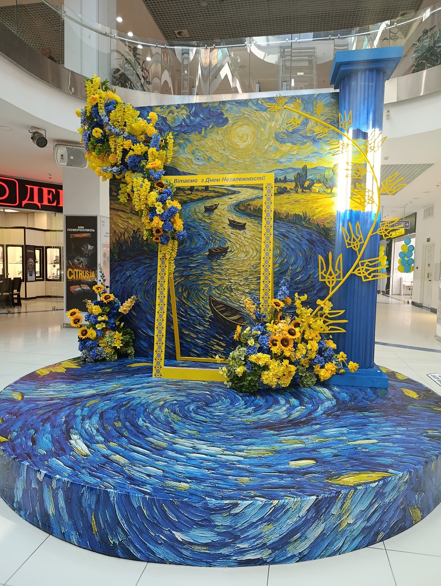 The image displays a festive installation within a shopping mall. The primary subject is a large, circular platform covered in a pattern resembling Vincent van Gogh's "The Starry Night," with swirling blues and yellows. In the center, a rectangular photo frame is positioned against a backdrop depicting a stylized Ukrainian landscape with a river and boats, also in a painterly style. The backdrop is adorned with blue and yellow floral arrangements. To the right, a tall blue pillar stands, decorated with yellow trident symbols, the national emblem of Ukraine. Yellow floral arrangements flank the photo frame and the base of the pillar. The overall presentation appears to be a decoration for a national holiday, likely Ukraine's Independence Day, given the prominent use of blue and yellow. In the background, the interior of a shopping mall is visible, with storefronts, escalators, and a railing on an upper level. No people are clearly visible in the foreground, though a person can be seen on the upper level of the mall. The lighting suggests an indoor environment with overhead illumination.