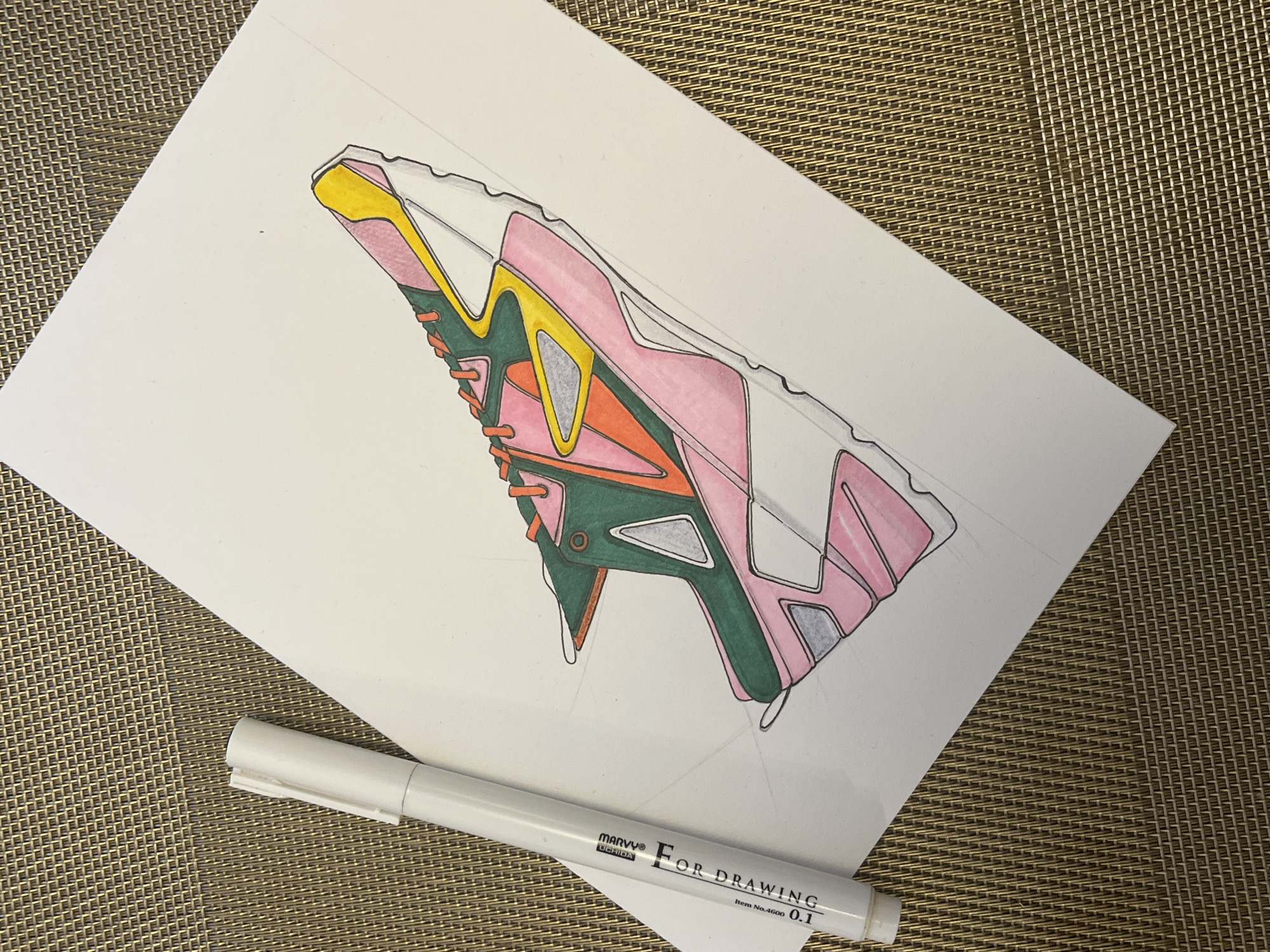 An angled overhead shot captures a detailed, colorful drawing of a futuristic or athletic shoe on a white sheet of paper. The drawing, seen from a side profile, features a dynamic design with panels colored in shades of light pink, vibrant yellow, dark green, bright orange, and light gray. The outlines of the shoe are crisp black lines, suggesting a fine-liner pen was used, and some sections, notably parts of the sole and upper, are still uncolored white, indicating the drawing is in progress.
The white paper lies diagonally across a textured, brown or golden-colored surface, which appears to be a woven mat or placemat. Resting partially on the paper and partially on the mat, below the drawing, is a white pen or marker. Visible text on the pen identifies it as "MARVY® Uchida" and states "FOR DRAWING Item No.4600 0.1", indicating it's a fine-tip drawing pen. The scene suggests a creative activity, possibly an artist's workspace where a shoe design is being developed or rendered.