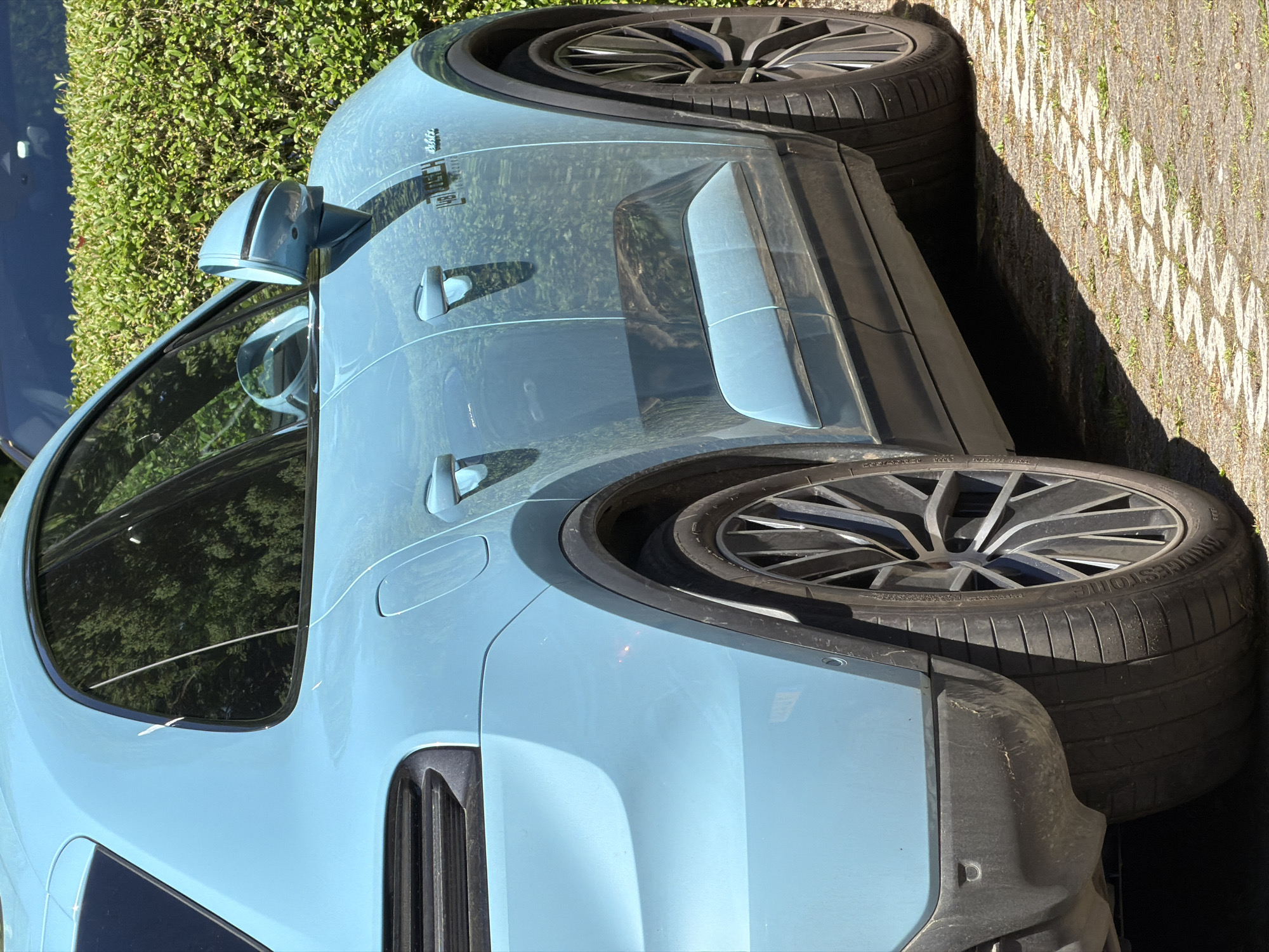 The image shows a light blue Porsche Taycan Cross Turismo parked on a cobblestone surface, likely in Fentange, Luxembourg. The focus is on the side of the car, highlighting its design elements like the door handles, side mirror, rear quarter panel vents, and the rear tire. The tires are Bridgestone. Green bushes are visible in the background, suggesting a residential or park area. There are no people visible in the image.