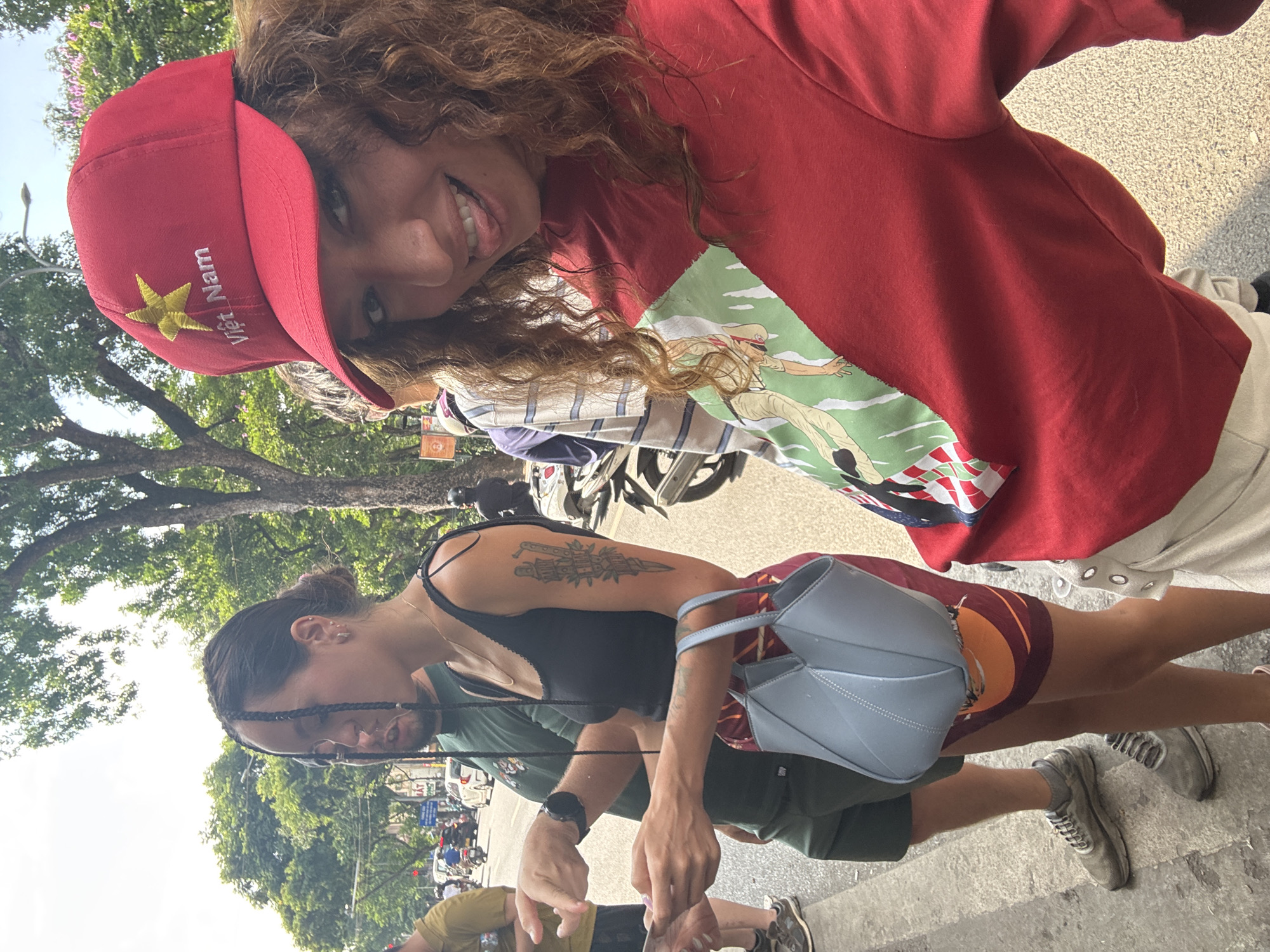 Three individuals are visible in the image. The primary subject is a young woman with curly brown hair, wearing a red sweatshirt and a red cap with a gold star and the words "Việt Nam." She is smiling at the camera.
Behind her, another woman with dark hair and a tattoo on her arm is wearing a black tank top and carrying a light blue purse. An additional individual, possibly male, is partially visible, wearing a green shirt.
The scene appears to be outdoors in Hà Nội, Vietnam. Motorcycles are parked in the background along a street lined with trees.