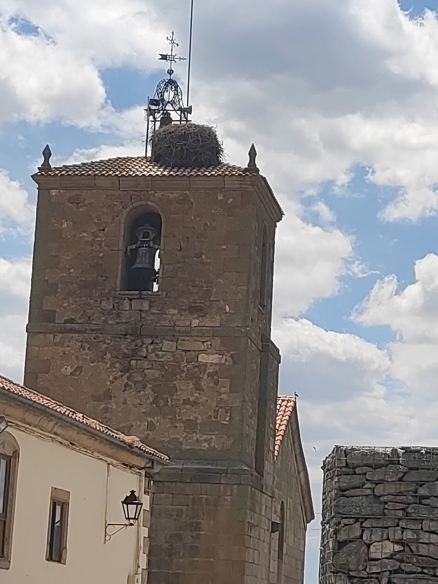 The image shows a close-up view of a stone bell tower in Sobradillo, Spain. The tower has a tiled roof and a bell visible through an arched opening. At the very top of the tower, a large nest, likely built by storks, is present, topped by a weather vane. The sky behind the tower is partly cloudy. In the foreground, to the left, a portion of a white building with a light fixture and a window is visible. To the right, a section of a stone wall constructed with gabions is also visible.