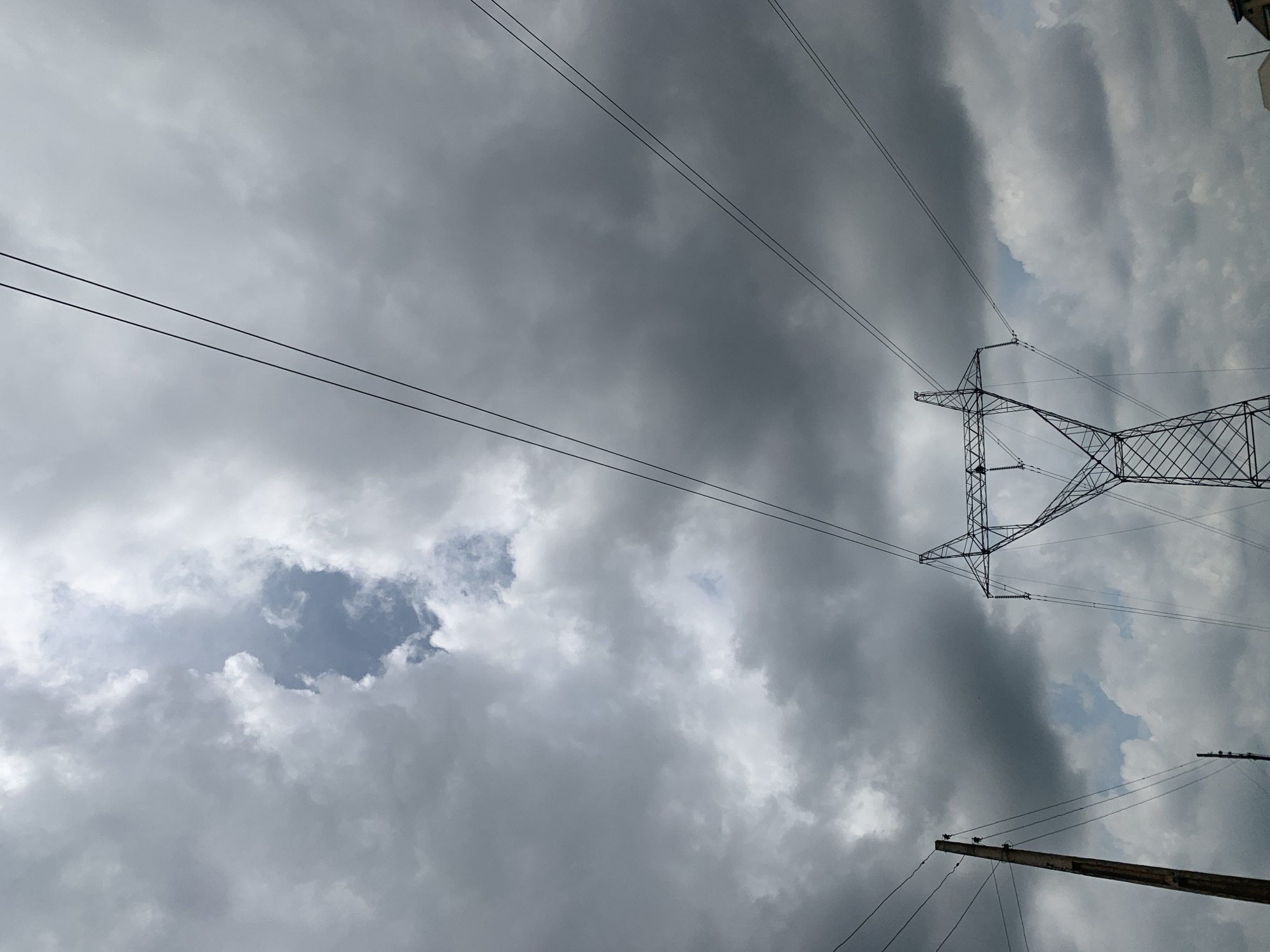 A low-angle shot captures a vast, overcast sky with dramatic, dark clouds. Two sets of power lines are visible, crisscrossing the frame. In the foreground, a tall, lattice-style electricity pylon dominates the right side of the image, its metallic structure stark against the cloudy backdrop. To the left, a wooden utility pole stands, also supporting power lines. The overall atmosphere is somber and somewhat foreboding due to the heavy cloud cover, suggesting an impending storm or a recent downpour. The scene is devoid of any human presence or immediate activity.