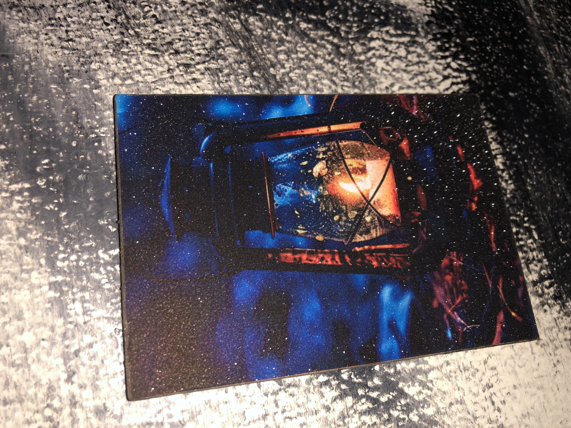 The image displays a close-up, angled shot of a printed image of a lit lantern against a dark, starry background. The lantern is old-fashioned, with a glass globe and a metal frame, and a flame is visible inside. The background is deep blue with many small white specks that resemble stars or glitter. The printed image is placed on a textured, gray and white surface. The scene does not depict any specific location within Kabong, Nigeria, nor does it contain any people or actions.