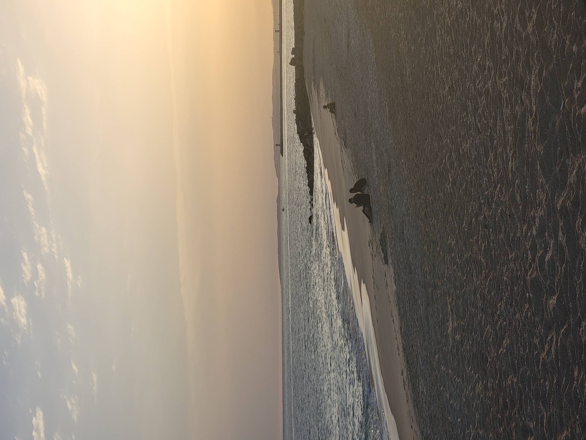 This image captures a serene sunset scene on the beach in Quarteira, Portugal. The sky transitions from a pale blue at the top to a warm, peachy-orange hue near the horizon, with scattered wispy clouds. The sea, a calm expanse of dark blue water, gently laps against the sandy shore.
In the foreground, the beach is textured with ripples from the receding tide and footprints. Several individuals are present:
* On the left, two figures are seated on the sand, silhouetted against the brighter background, seemingly engaged in conversation or resting.
* Further to the right, another solitary figure sits on the sand, also in silhouette.
* In the distance, a long, rocky breakwater extends into the sea, dotted with small structures that appear to be lighthouses or navigation markers. The coastline stretches along the horizon, with hints of buildings and structures suggesting the presence of the town.
The overall atmosphere is peaceful and tranquil, characteristic of a late afternoon or early evening on a Portuguese beach.