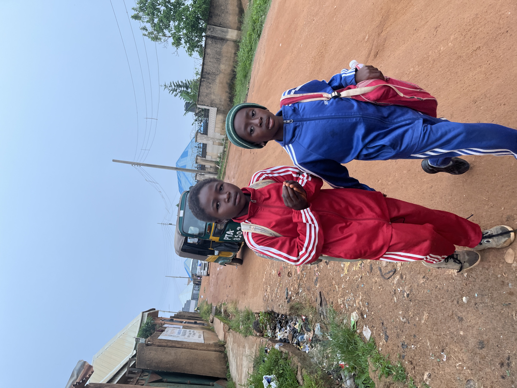 Two young children walk down a dirt road in Jos, Nigeria. The child on the left wears a red tracksuit with white stripes and carries a backpack. The child on the right wears a blue tracksuit with white stripes, a green beanie, and a red and white bag. In the background, a yellow and green auto-rickshaw, also known as a "keke napep", is parked on the side of the road. The environment appears to be an urban or semi-urban setting, with buildings and vegetation visible along the roadside. There is a drain alongside the road filled with debris and waste, and a power pole with electrical wires is present. The sky is overcast.