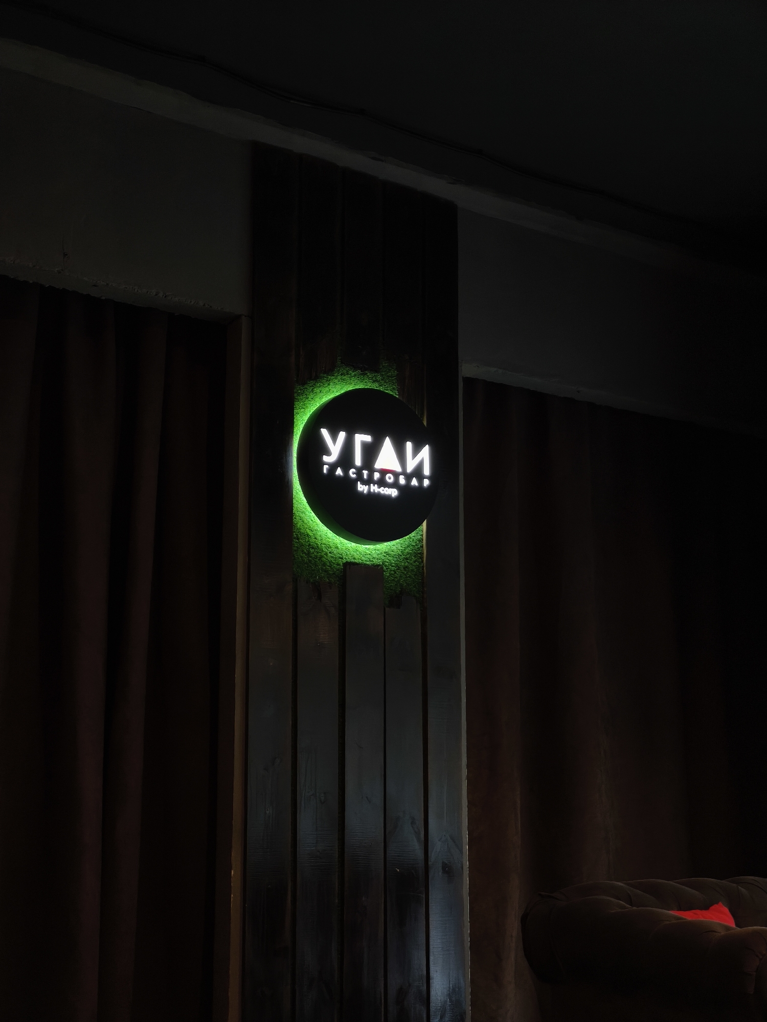 The image captures an indoor, dimly lit scene, likely an establishment such as a bar or lounge. The central subject is a circular sign featuring white Cyrillic text, illuminated by a bright green glow that creates a halo effect around its edge. The text on the sign reads "УГАИ" (UGAI) in large letters, with "ГАСТРОБАР" (GASTROBAR) below it, and "by H-corp" in smaller Latin script at the bottom.
The sign is mounted on a dark wooden slatted wall panel. To the left and right of this panel, dark brown, heavy curtains frame the scene, contributing to the muted and intimate atmosphere. In the bottom right corner, a portion of a dark brown, tufted sofa or armchair is visible, with a small, bright red object (possibly a cushion or a design detail) providing a stark color contrast.
The low lighting suggests it is evening or night. There are no people visible, and no specific activity is taking place; the focus is on the interior decor and signage. The overall impression is one of a modern and cozy venue, consistent with a gastrobar. The location context is Chaikovsky, Russia.