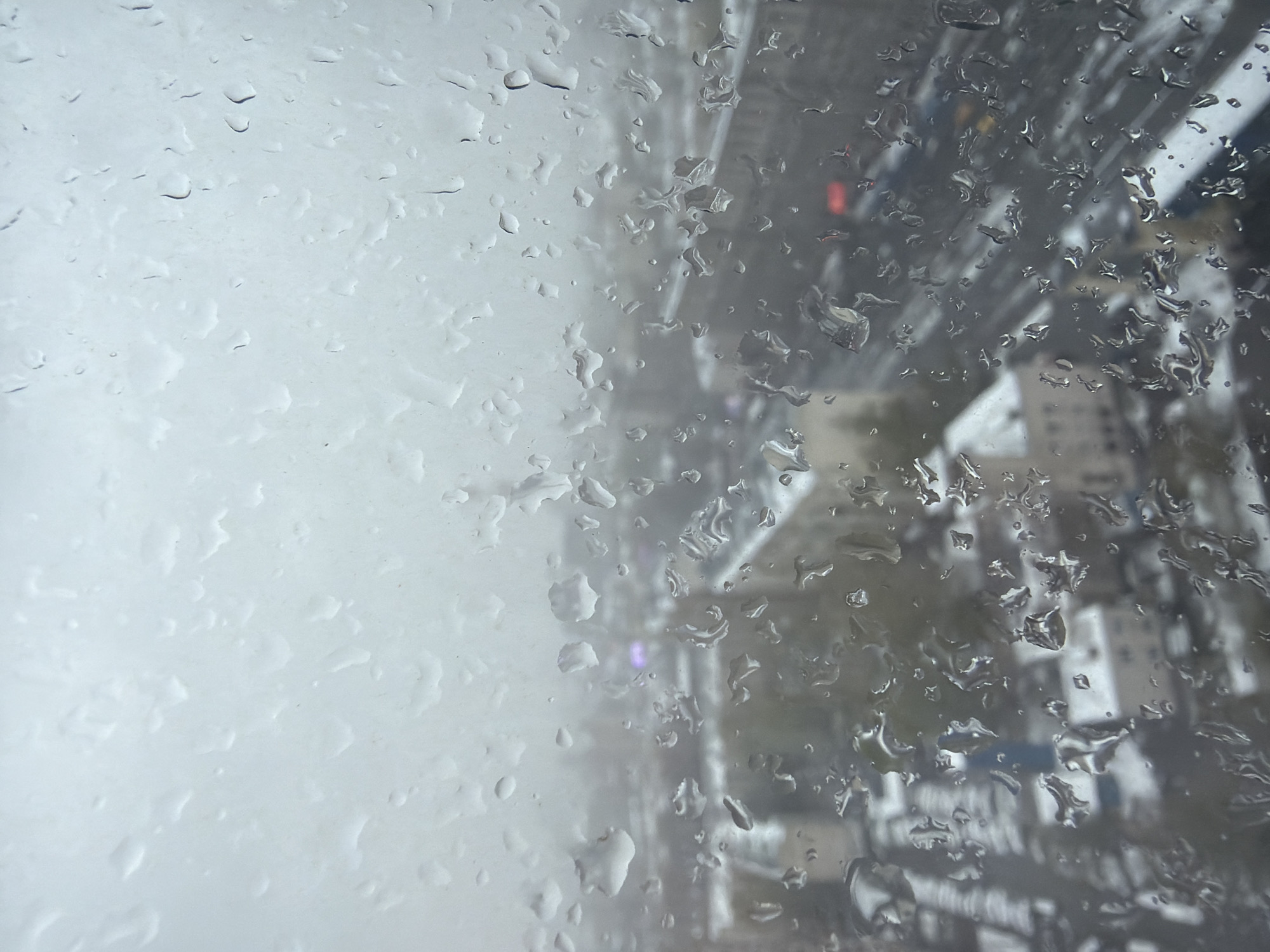 The image captures a close-up view of a transparent surface, likely a window, extensively covered in raindrops and water streaks. The left side of the frame is lighter and more diffused, dominated by smaller, less distinct water droplets that create a soft, textured appearance. Towards the right, the raindrops are larger and more numerous, causing greater distortion of the background.
Through the rain-obscured glass, a blurred urban landscape is visible. Indistinct shapes of buildings and dark, linear structures, possibly an elevated roadway or bridge, can be faintly discerned. Scattered points of artificial light pierce through the blur, notably a prominent red glow, likely from a traffic light or vehicle, and a fainter purple or pink light in the middle-right section.
The scene depicts an observation point from an interior space looking out onto a city during rainy conditions. The time of day appears to be evening or night, suggested by the visible artificial lights against the dim, urban backdrop. No specific objects or individuals are clearly identifiable in the heavily blurred background, and no text is visible. The general setting is consistent with a large urban environment such as Moscow, Russia.