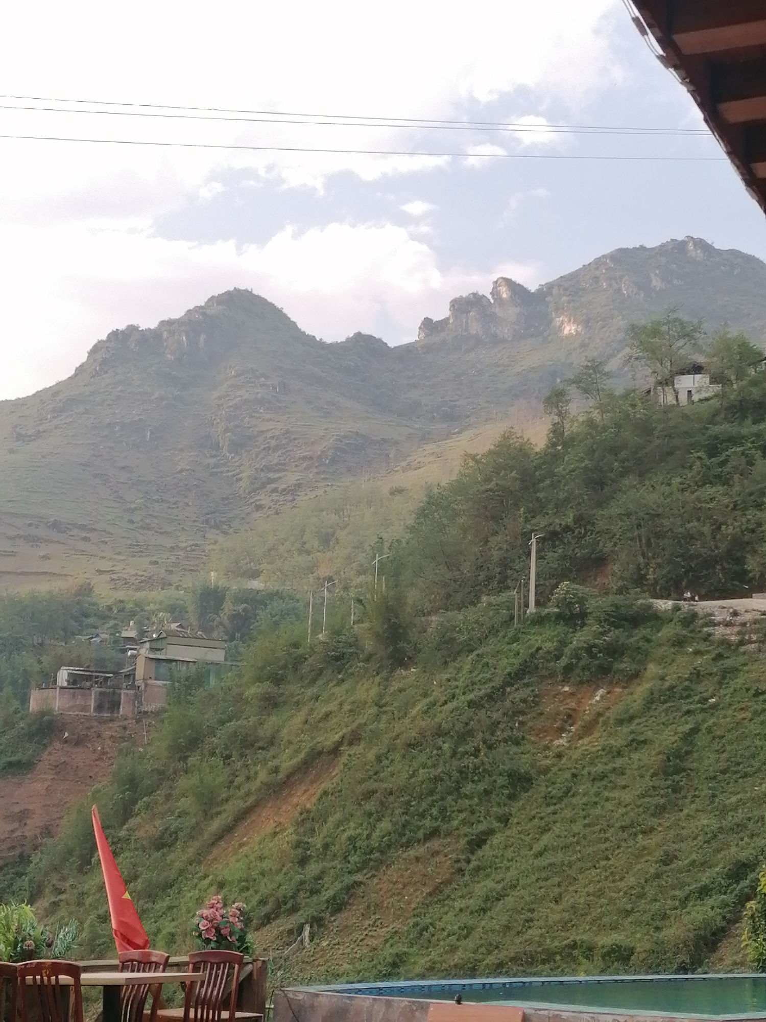 This image captures a panoramic view of a lush, mountainous landscape, consistent with the terrain found in Sa Pa Ward, Vietnam. Verdant mountains, densely covered in green foliage, dominate the background, displaying rugged peaks and rocky outcrops under a partly cloudy, daylight sky.
In the foreground, an outdoor seating area is visible on the left, featuring a wooden table and chairs, and a pot of pink flowers. A prominent red Vietnamese flag with a yellow star is planted nearby, indicating the national identity of the location. To the right, the bright blue water of a swimming pool is partially visible, suggesting a resort or guesthouse setting.
The middle ground and mid-slopes of the mountains are dotted with various buildings and infrastructure. Several structures with metal roofs and concrete bases are nestled into the hillside, alongside a smaller, white house perched higher up. Utility poles and power lines crisscross the scene, connecting these developments. A winding road or path is also discernible on the hillside, with what appear to be two small, blurry figures on it. The overall impression is one of a serene natural environment where human habitation is integrated into the mountainous terrain. No specific activity is clearly depicted, and no legible text is visible other than the emblem on the flag. The lighting suggests it is daytime, possibly late morning or afternoon, with soft, diffused light from the overcast sky.