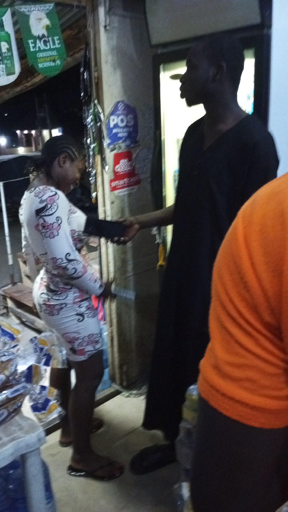 Outside a shop in Adankolo, Nigeria, a man and a woman are shaking hands. The woman wears a floral-patterned dress and sandals, while the man wears a long black shirt and black shoes. A person in an orange shirt stands to the right of the frame. POS and smartcash posters are on the wall beside them. An "EAGLE" brand schnapps sign hangs overhead, and packaged goods are on a shelf in the foreground.