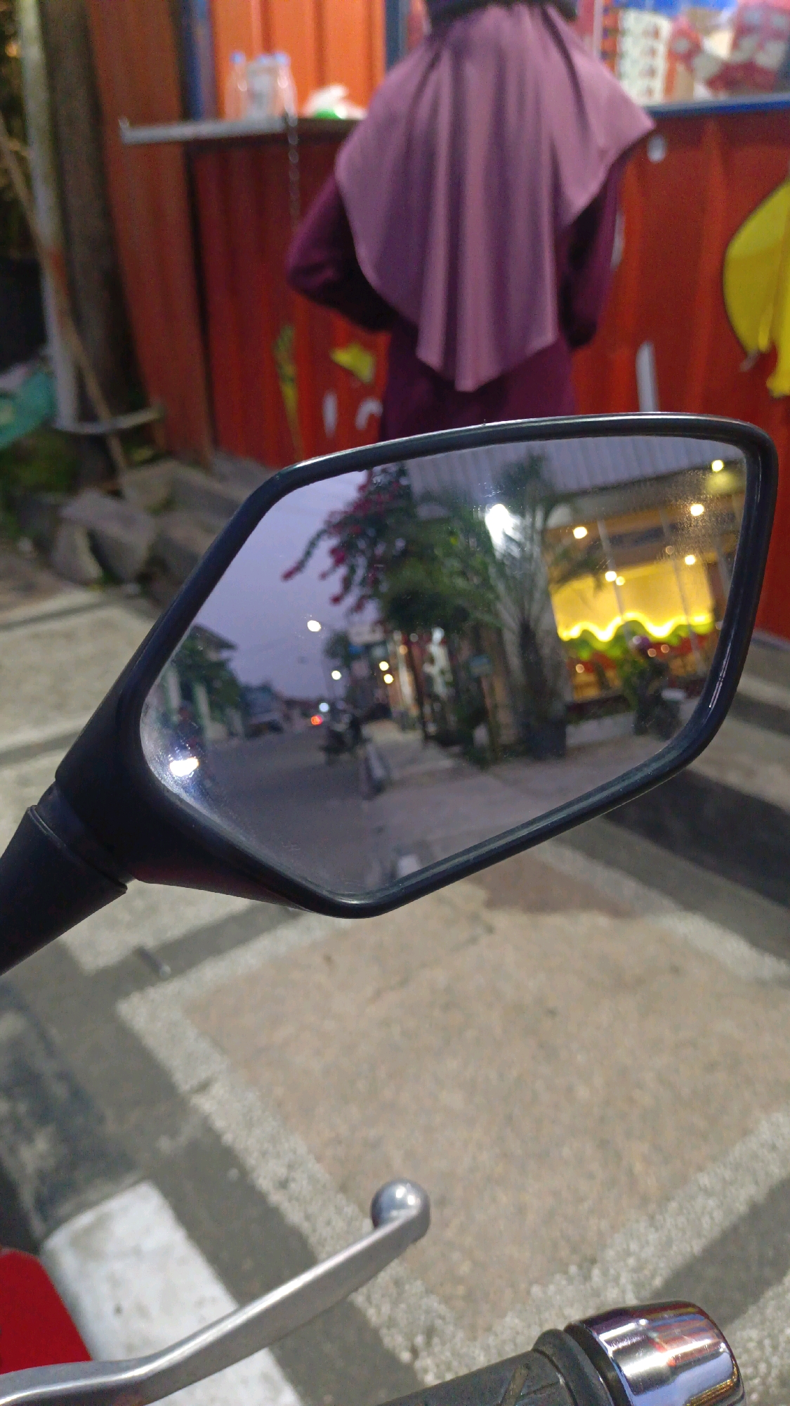 The image captures a reflection in a motorcycle's side mirror, showing a street scene in Padang Panjang, Indonesia. The reflection includes buildings with illuminated yellow awnings, streetlights, trees, and a motorcycle. In the foreground of the main image, the handlebars and brake lever of a motorcycle are visible, along with a person wearing a purple hijab and jacket standing in front of a red stall. The ground is paved with concrete blocks. The lighting suggests it is dusk or evening.